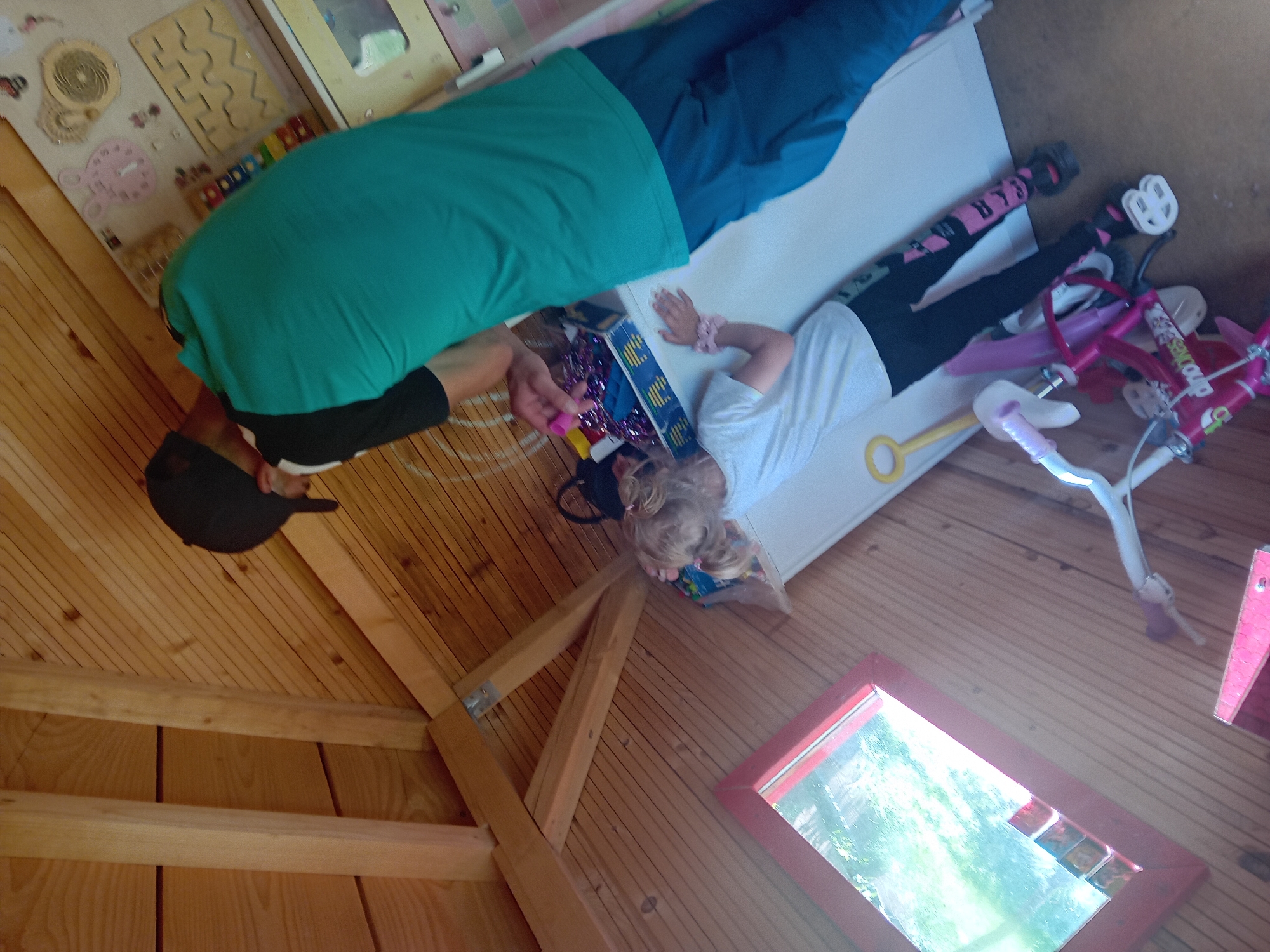 In Zuberec, Slovakia, a young blonde-haired girl wearing a white t-shirt and black leggings with pink lettering is lying down in a wooden structure that appears to be a playhouse. She is positioned next to a small pink bicycle. A person, whose face is obscured by a black baseball cap, is standing over the girl, wearing a teal t-shirt and blue pants. The person's right arm is extended downwards, reaching towards the girl. The interior of the wooden structure features a red-framed window reflecting greenery and trees. A busy board with various toys and activities is visible in the background, mounted on a white wall. The floor is made of wooden planks.