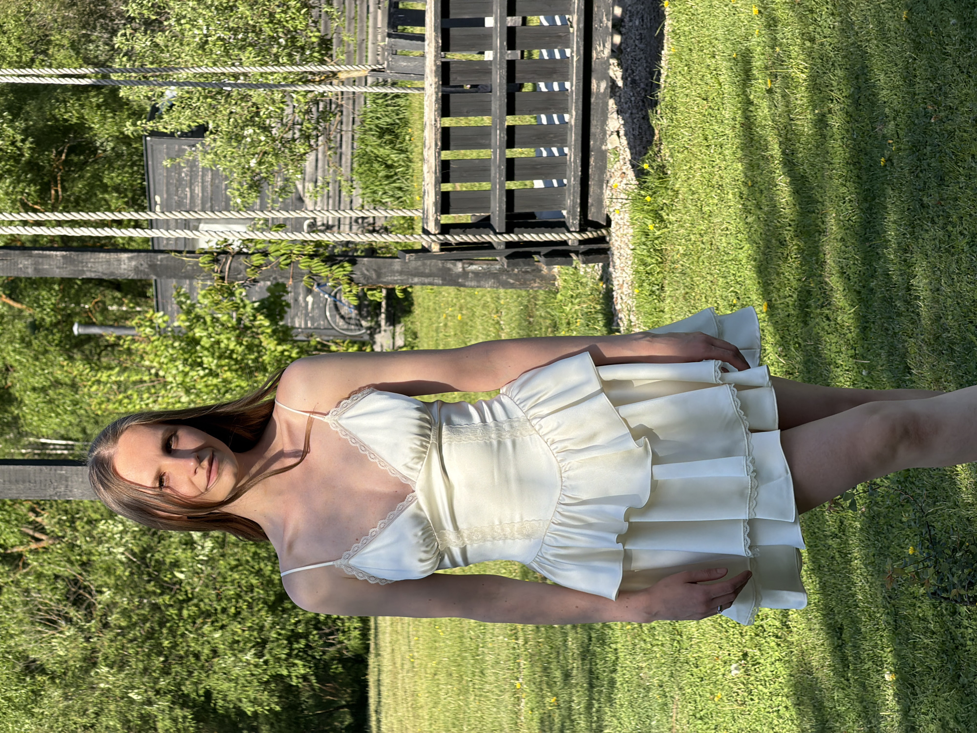 A woman stands on a lawn in Prudki, Russia. She wears a light-colored dress with lace trim and spaghetti straps. The lawn is green, with some yellow flowers. Behind her is a black wooden structure, and further in the background are trees and foliage. The scene is outdoors in daylight.