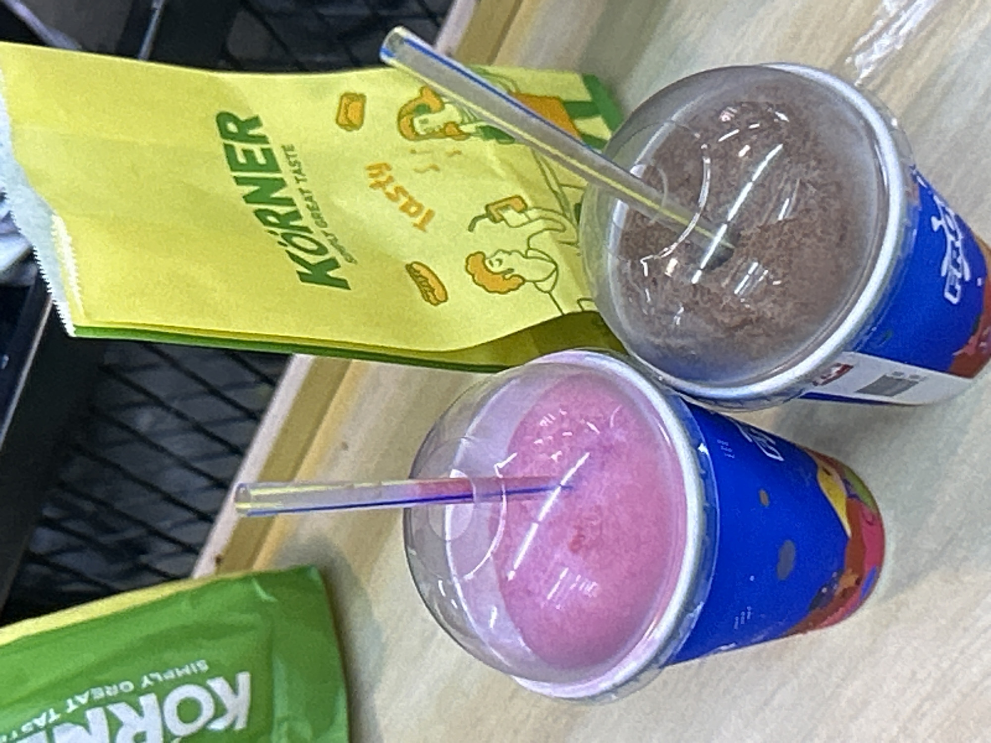 Two slushy drinks, one pink and one chocolate, are placed on a light brown wooden surface. Both drinks are in blue cups with clear plastic lids and colorful graphics on the cups. Each drink has a straw inserted. To the right of the drinks is a yellow paper bag with the brand name "KÖRNER" printed in green. The bag also features cartoon drawings of people and food items, along with the text "SIMPLY GREAT TASTE" and "Tasty." In the background, a green bag with white text, also appearing to be from the same brand, is partially visible. The scene appears to be indoors, possibly at a food stall or convenience store in Depok, Indonesia. The lighting is bright, creating reflections on the plastic lids of the drinks.