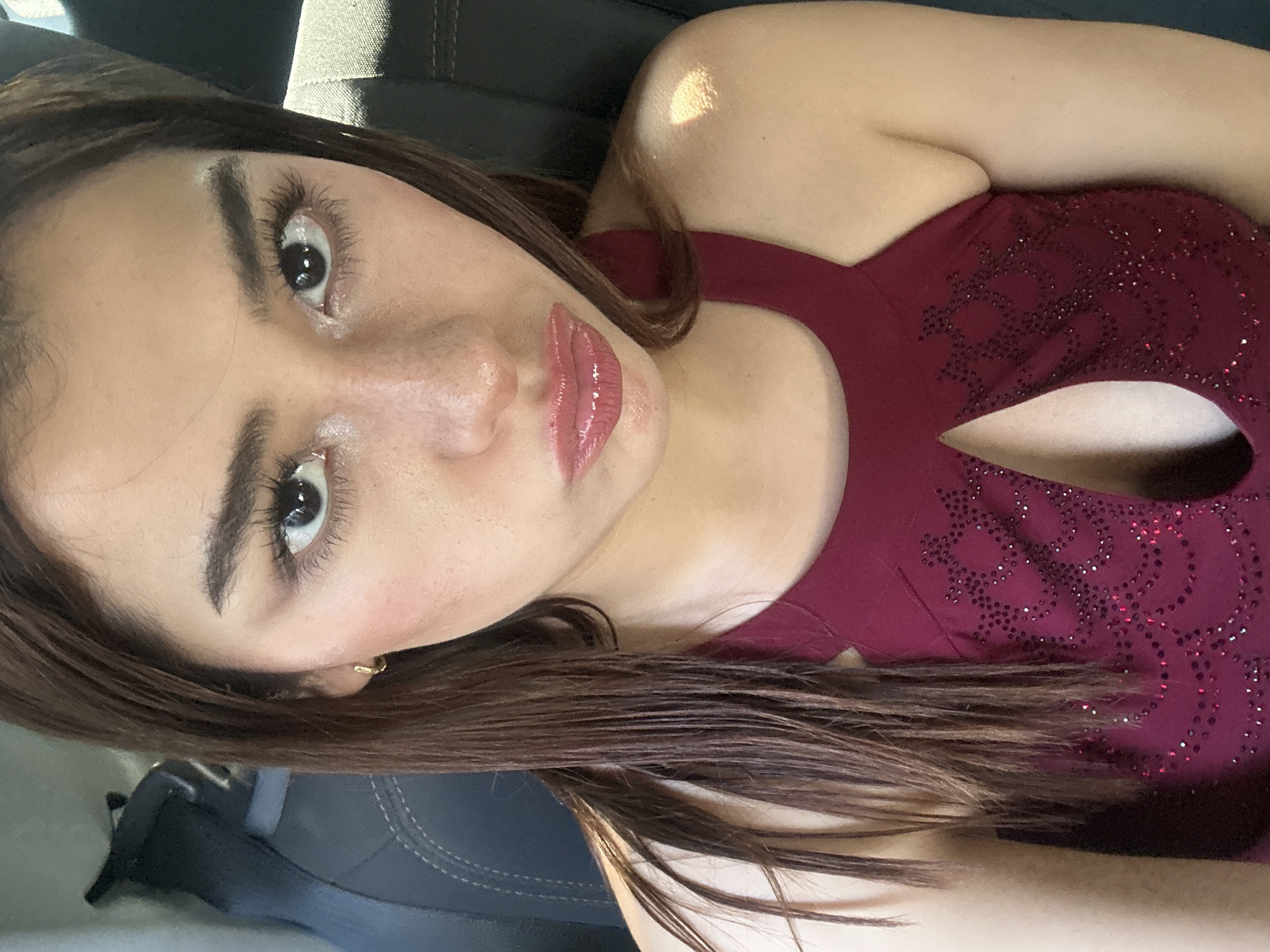 A woman with brown hair is wearing a maroon dress with a cutout in the chest and is looking at the camera. The woman appears to be in a car. No location information is available.