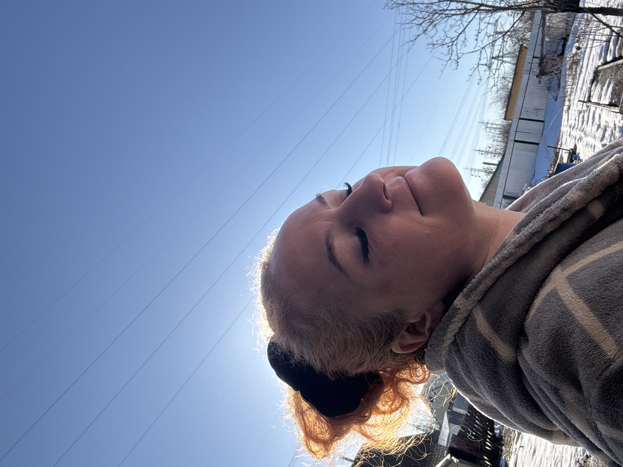 The image is a low-angle shot of a person with their eyes closed, appearing to be looking up at the sky. The person has fair skin, light-colored hair with darker roots and a black hair tie, and is wearing a gray and white striped robe. The sky is a clear blue with several visible power lines running across it. The sun is positioned behind the person's head, creating a bright halo effect around their hair. In the background, there is a view of what appears to be a rural area with some snow-covered ground, bare trees, and buildings. The buildings appear to be simple, possibly garages or sheds, and are partially obscured by the angle and distance. The specific location in Chaikovsky, Russia is not determinable from this photograph.