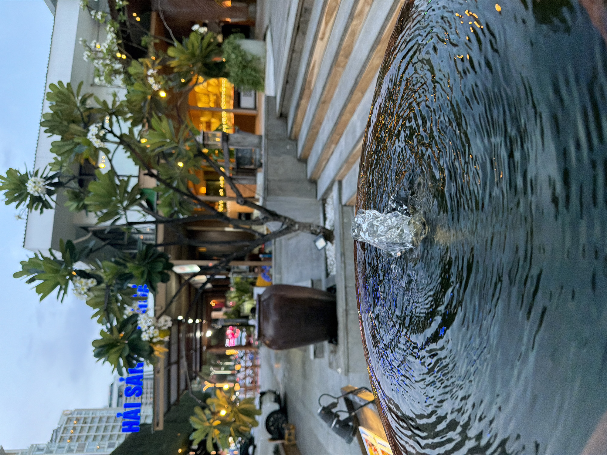 The image shows a close-up view of a water fountain, with ripples on the surface of the water. In the background, a street scene is visible, including buildings, trees, and a large ceramic pot. There is a sign with blue lettering that appears to read "HẢI SẢN," which translates to "seafood" in Vietnamese, suggesting the location is a seafood restaurant or market. The overall lighting suggests it is either early morning or late evening. This scene is located in Nha Trang Ward, Vietnam, a coastal city known for its beaches and seafood restaurants.