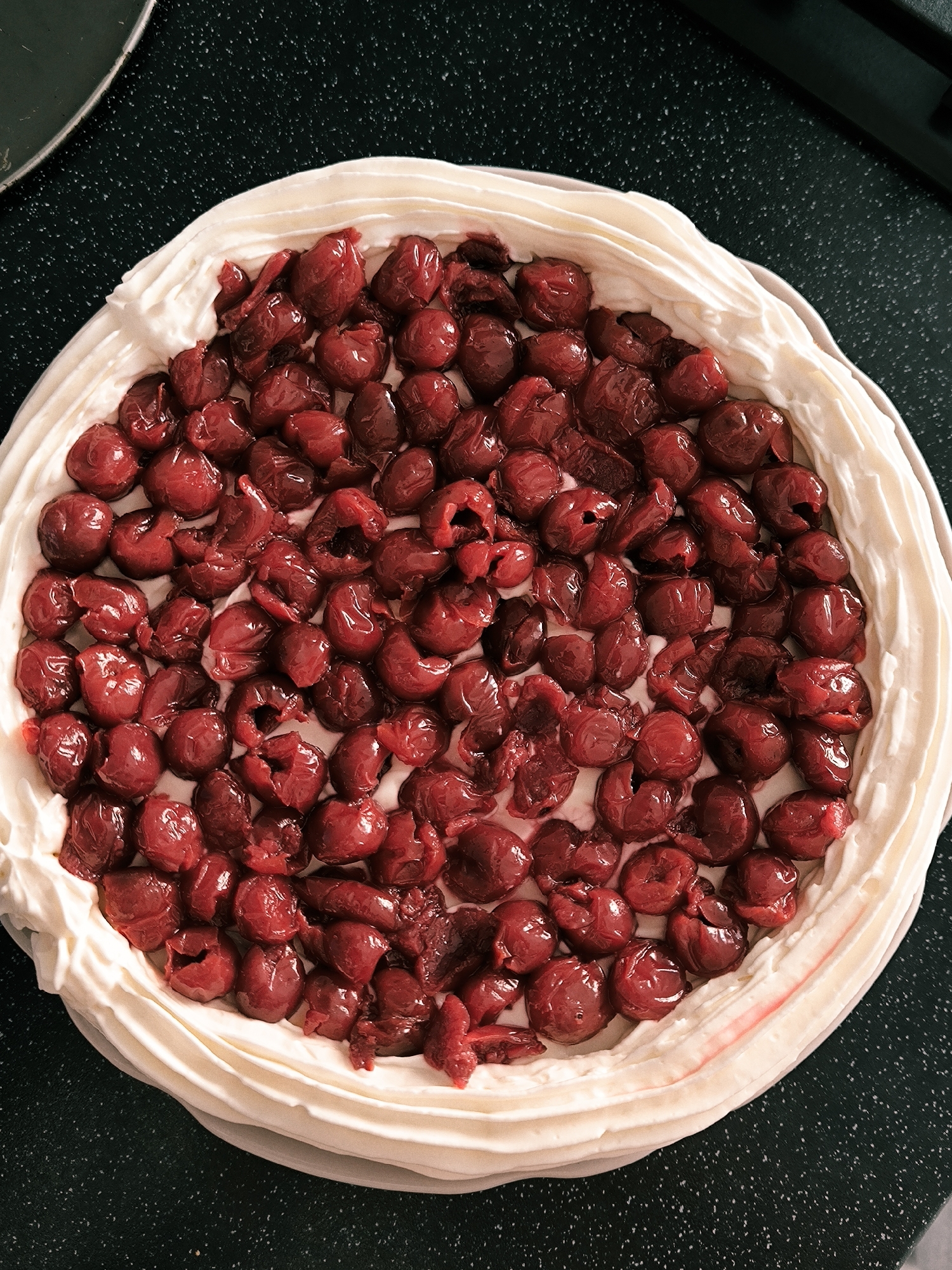 The image displays a close-up, overhead view of a round dessert. A dense layer of dark red cherries, mostly pitted and some appearing split, occupies the center of the dessert. This cherry layer rests on a base of white or off-white cream, which is piped in undulating, concentric ridges around the dessert's perimeter. Minor reddish discoloration from cherry juice is visible on sections of the cream. The dessert is positioned on a dark, speckled surface, consistent with a kitchen countertop. A partial, curved edge of a dull greenish-grey object is visible in the upper-left quadrant of the frame. No individuals are depicted. The specific location within Vinnytsia, Ukraine, cannot be determined from the visual evidence provided.