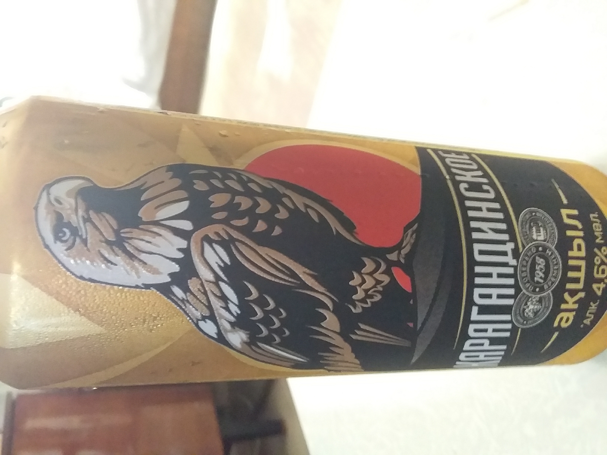 A close-up shot shows a can of "KARAGANDINSKOE AKZHUL" beer, featuring an illustration of a hawk. The can is primarily gold and black with a red circular emblem. Text on the can reads "1958" and "АЛК. 4,6% МВЛ". The can is covered in condensation, indicating it is cold. The background is blurred, with a wooden surface visible on the left and a white surface on the right. The can is likely a product of Kazakhstan, given the name "Karaganda," a major city in that country. There are no people or any indication of location within the city of Saran or its administration.