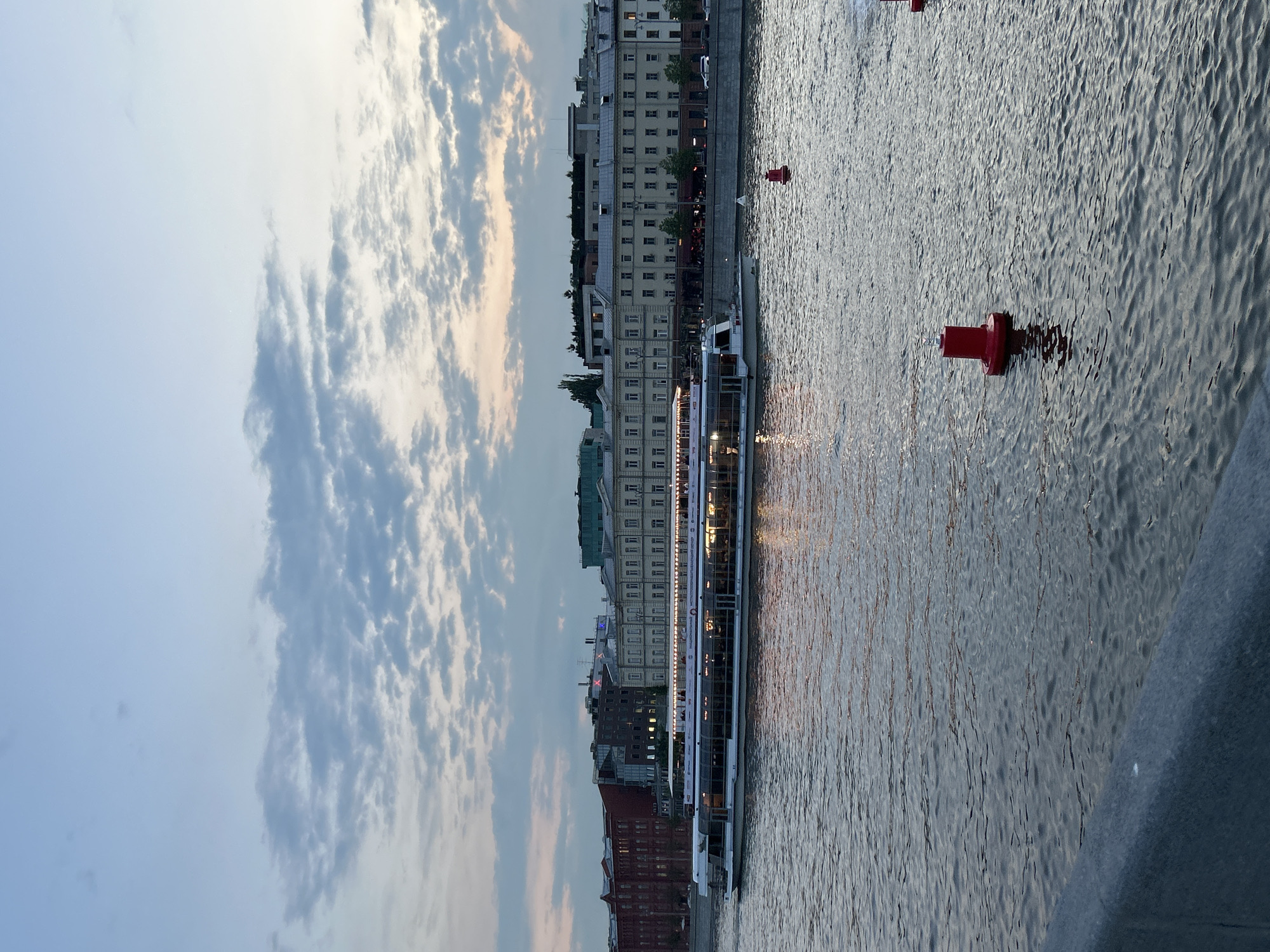 The image shows a body of water, possibly a river, with a large passenger boat running along its length. The boat is flanked by buildings on one side, displaying a range of architectural styles and colors, suggesting an urban environment. Red buoys are visible in the water, marking a channel or navigational path. The sky is partially cloudy, with varying shades of blue and white, indicating either early morning or late afternoon. The environment seems peaceful and suggests a city setting.