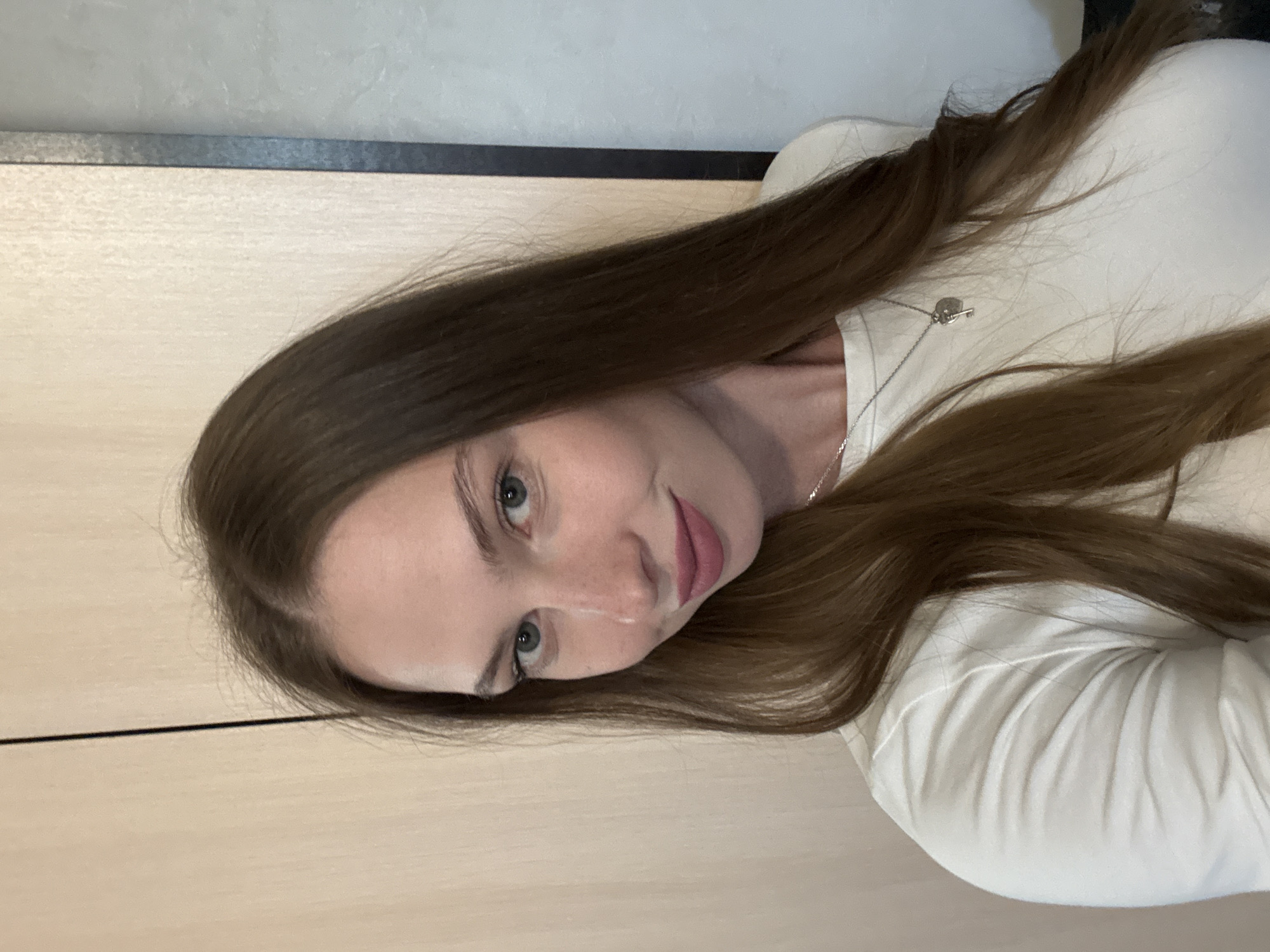 A young woman with long, brown hair and blue eyes is posing for a selfie. She is wearing a cream-colored long-sleeved shirt and a silver necklace with a key pendant. She has freckles on her nose and cheeks and is wearing pink lipstick. The background appears to be a neutral-colored wall with a vertical seam. The lighting is soft and natural. The overall impression is casual and relaxed. The image is taken indoors. This image cannot be linked to a specific location in the city of Perm, Russia.