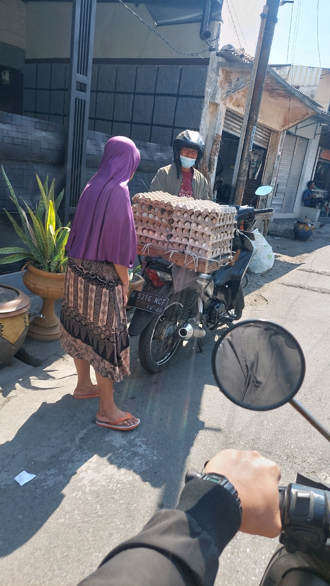 In Sidoarjo, Indonesia, a person wearing a purple hijab and a patterned skirt is standing next to a motorcycle loaded with egg cartons. The person is facing away from the camera, looking at another individual who is wearing a helmet and a blue mask. This second person appears to be the driver of the motorcycle and is looking towards the person with the hijab. The motorcycle is black and has a license plate that reads "W 5316 NCF". In the background, there are buildings, a utility pole, and a plant in a pot. The scene is lit by sunlight, casting shadows on the ground.