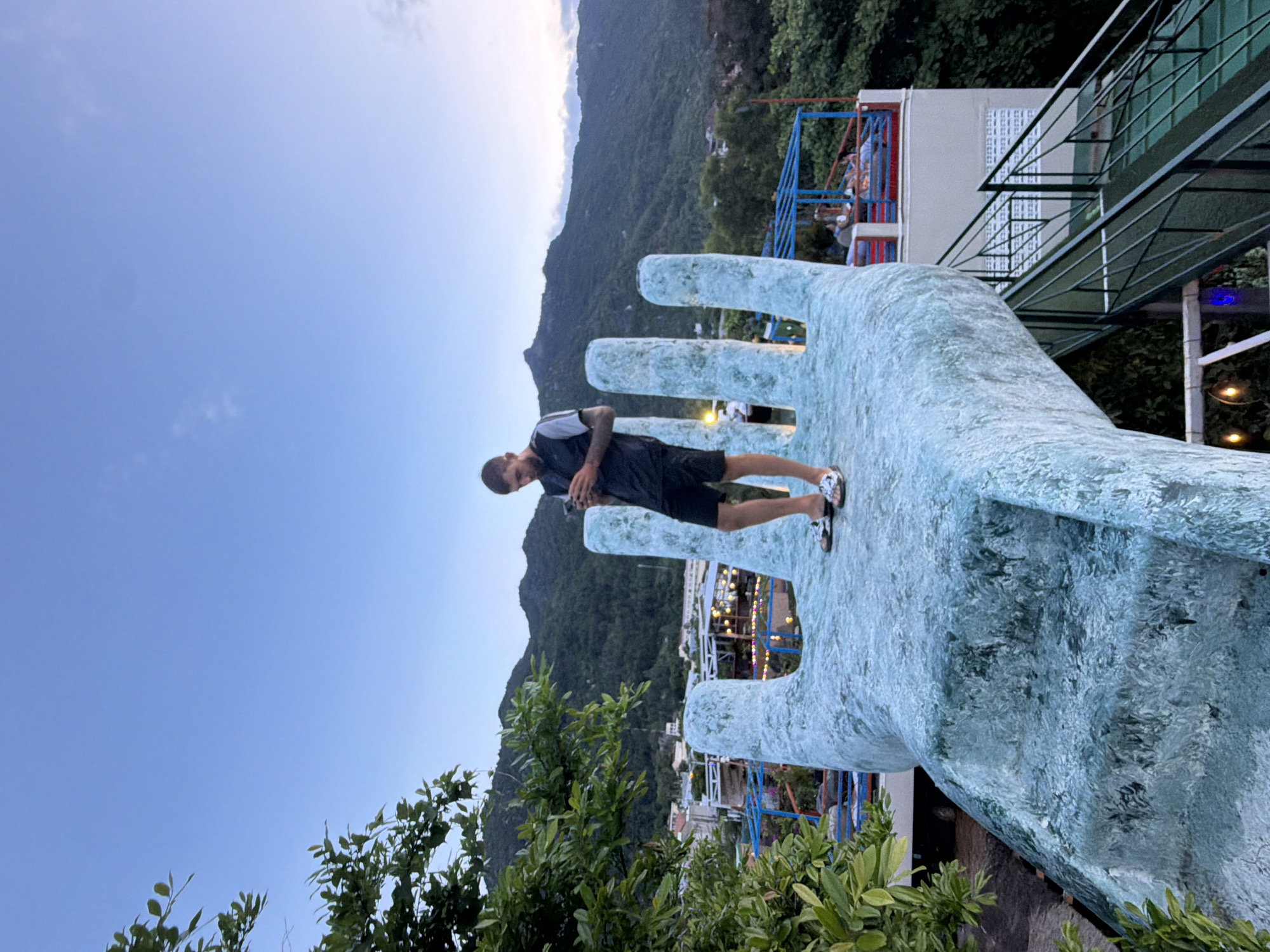 A young man stands on a large, pale blue, textured sculpture of a hand, holding his phone and looking down at it. He is wearing a dark grey vest over a black t-shirt, black shorts, and flip-flops. The hand sculpture is part of an outdoor setting, with a lush green hillside and mountains in the background under a clear, pale blue sky. In the middle ground, buildings with red and blue railings and some illuminated lights are visible. There are also trees and foliage in the foreground and to the left. A metal staircase with a green surface is visible to the right of the hand sculpture. The scene appears to be in the city of Nam Nha Trang Ward, Vietnam.