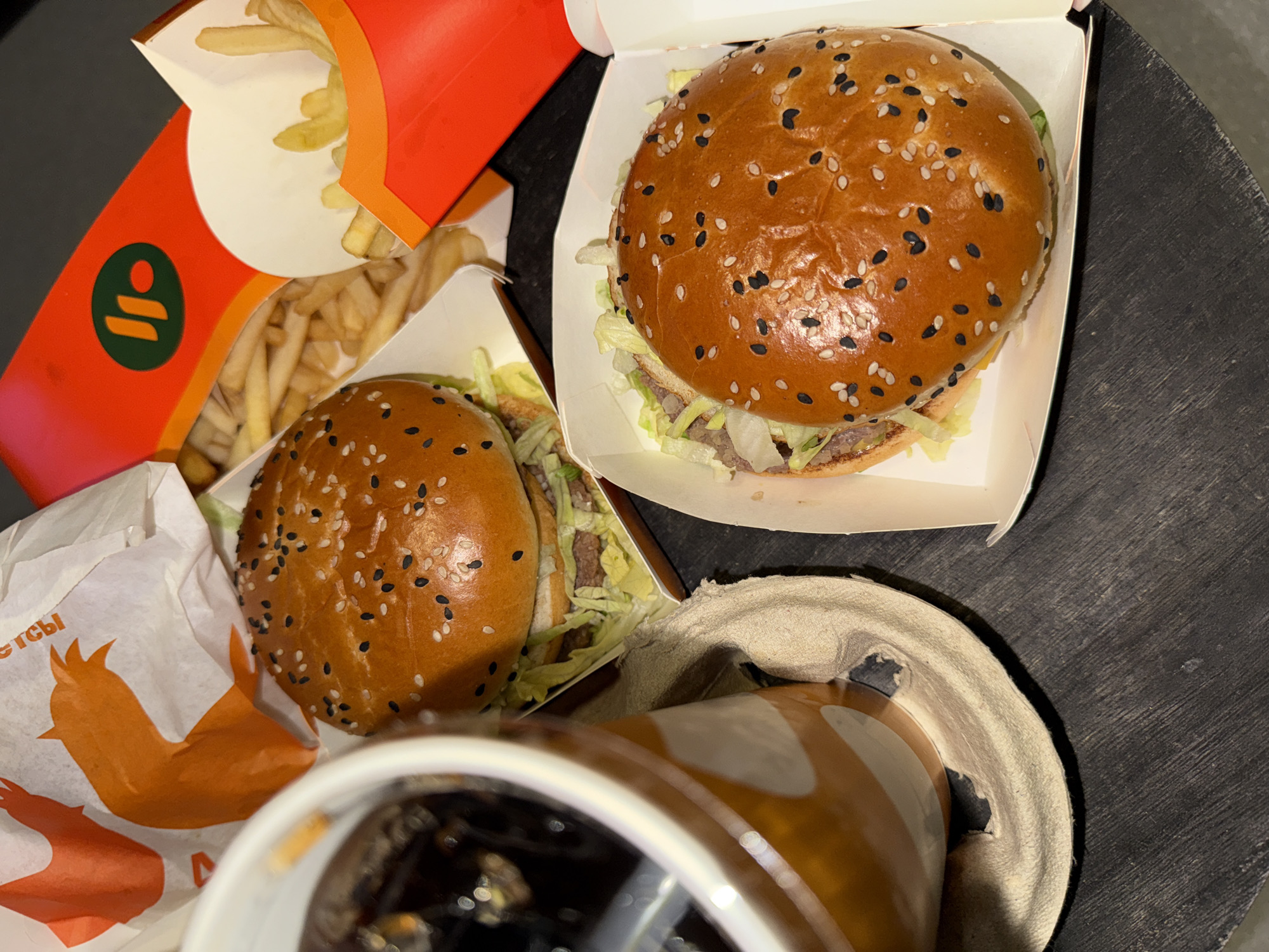 A still, top-down shot shows a fast-food meal on a black surface. The meal includes two burgers in cardboard containers, one with an orange wrapper featuring an abstract chicken wing and Cyrillic text. A red container holds French fries, with the restaurant's logo visible. A partially obscured soda cup sits in a tan cardboard holder. The location is identified as Perm, Russia, based on the Cyrillic text. The focus is on the arrangement of food, suggesting a meal setting.