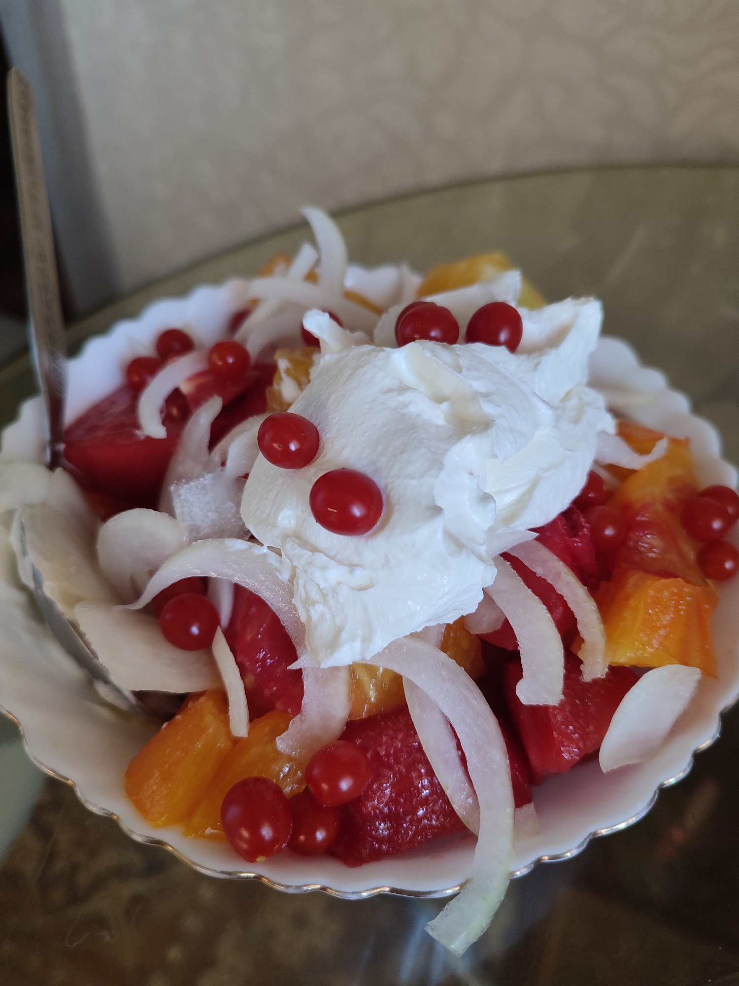 A close-up, eye-level, outdoor, full shot shows a white, scalloped-edged bowl filled with a fruit salad. The salad consists of chopped red fruit, such as strawberries or raspberries, and orange fruit, possibly cantaloupe or oranges. It is topped with a dollop of whipped cream and garnished with small, red berries, possibly cranberries or red currants. Thin, white slices, resembling onion or jicama, are scattered throughout the salad, adding an unusual textural contrast. A silver spoon is visible on the left side of the bowl, partially submerged in the fruit. The bowl rests on a dark, reflective surface, likely a table, in what appears to be an indoor setting with a neutral-colored wall in the background. The lighting is soft and natural, highlighting the vibrant colors of the fruit and the creamy texture of the whipped cream.