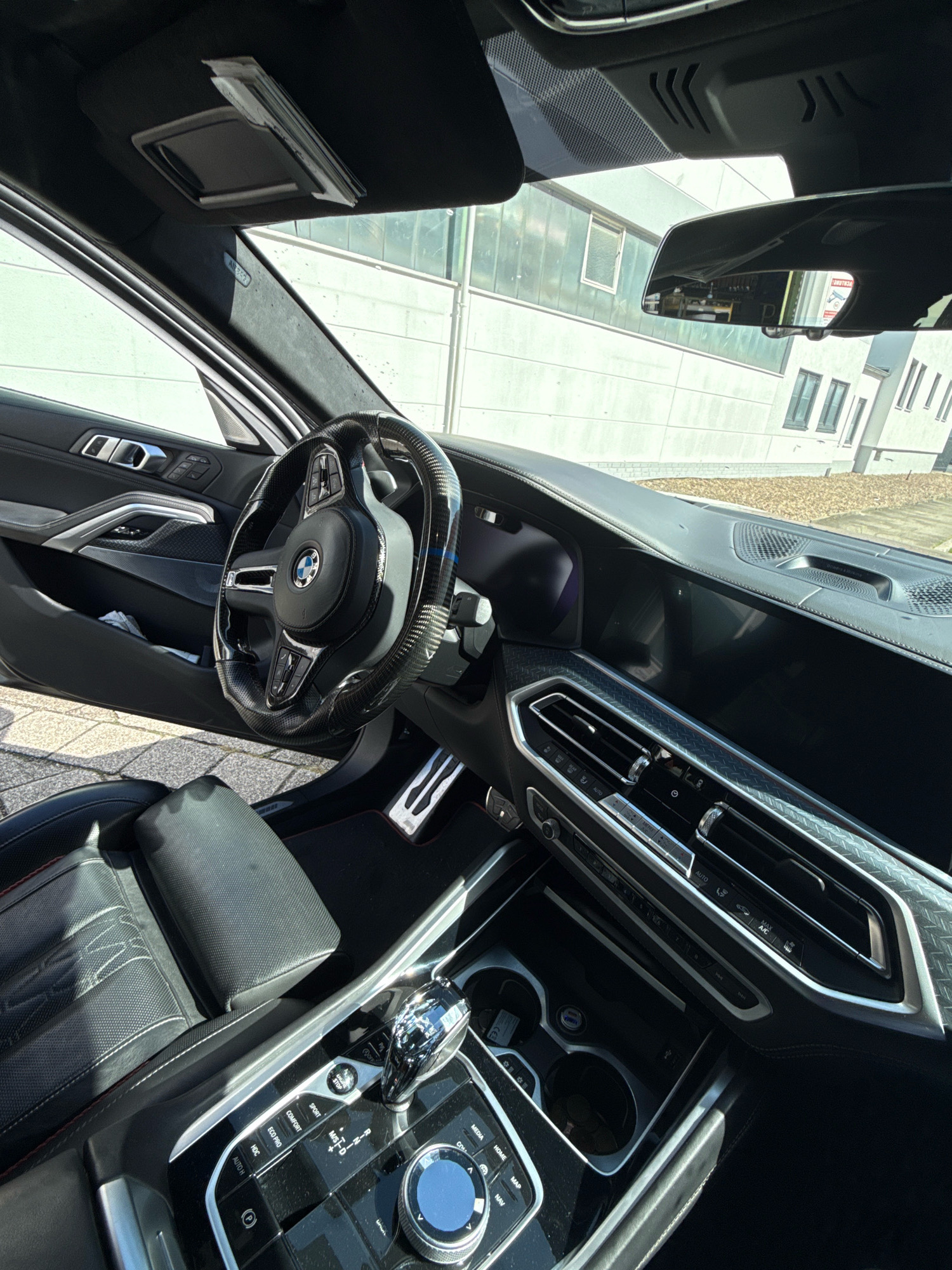 This is a first-person perspective, eye-level interior shot of a modern black BMW, likely taken in a parking lot. The car's black leather interior is visible, featuring a carbon fiber steering wheel with the BMW logo, a digital dashboard, and a central console with climate controls and a gear shifter. Sunlight streams into the cabin, highlighting the dashboard and the driver's side door. Outside the car, a building with light-colored siding and windows is visible, suggesting an urban or suburban setting. The ground is paved with cobblestones. No people are present in the image. The license plate is not visible. The reflection in the rearview mirror shows a building exterior, including windows and a parked vehicle. The scene appears to be in Bremen, Germany, based on the style of the building and the car's interior.