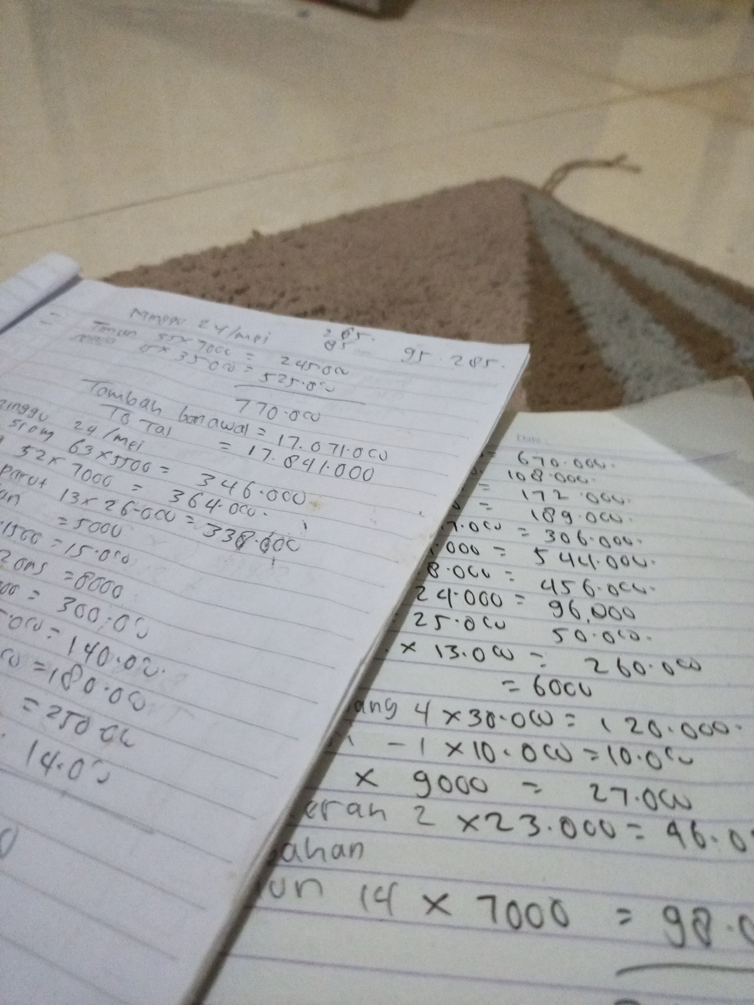 The image depicts a close-up, overhead view of a notebook filled with handwritten calculations and some loose papers with similar content, all resting on a patterned rug. The lighting suggests an indoor setting, potentially a home or office environment. The notebook pages are lined, and the handwriting is in Indonesian, featuring numbers, mathematical symbols, and some words. The calculations appear to be financial or accounting-related, with line items that include dates like "24/mei" (May 24th) and figures such as "7000," "3500," and larger sums involving commas as thousands separators.
On one page, there's a heading "Jumlah bdnawal" which translates to "Initial total." Another section includes "To Tal" and "Tg Tal" which likely refer to "Total" and "Total Tagihan" (Total Bill) respectively. Various multiplications and additions are evident, for example, "63 x 5500 = 346.000" and "13 x 26.000 = 338.000."
The loose papers to the right of the notebook also contain calculations. One entry is "670.000." Other lines show operations like "4 x 30.000 = 120.000," "9000 = 27.000," and "2 x 23.000 = 46.0". A prominent line at the bottom reads "14 x 7000 = 98."
The environment is not explicitly identifiable as Lhokseumawe, Indonesia, beyond the use of the Indonesian language in the notes. The rug has a distinct geometric pattern of alternating dark and light stripes, creating a diagonal effect. There are no people visible in the image, and the focus is entirely on the written documents and the surface they are placed on. The overall impression is one of a private workspace where financial records are being meticulously kept.