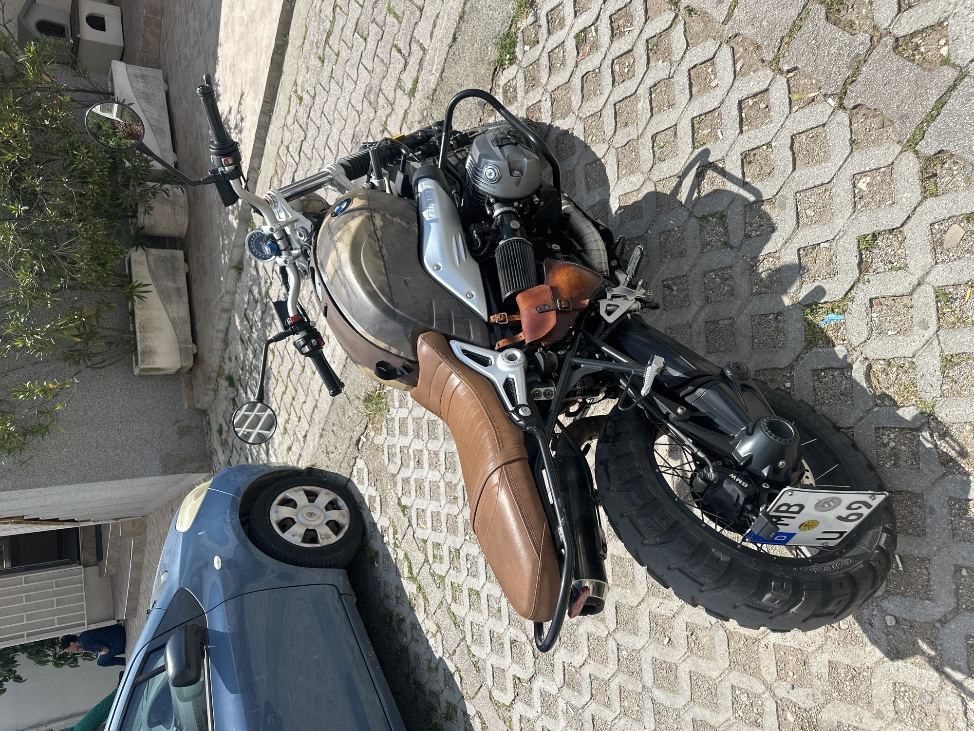 A parked BMW R nineT Scrambler motorcycle, with license plate "MB U 69", sits on a paved surface. A blue Toyota Yaris is parked behind the motorcycle. The setting appears to be an outdoor urban area, likely a street, in Budva, Montenegro. The photograph is taken from a high-angle perspective.