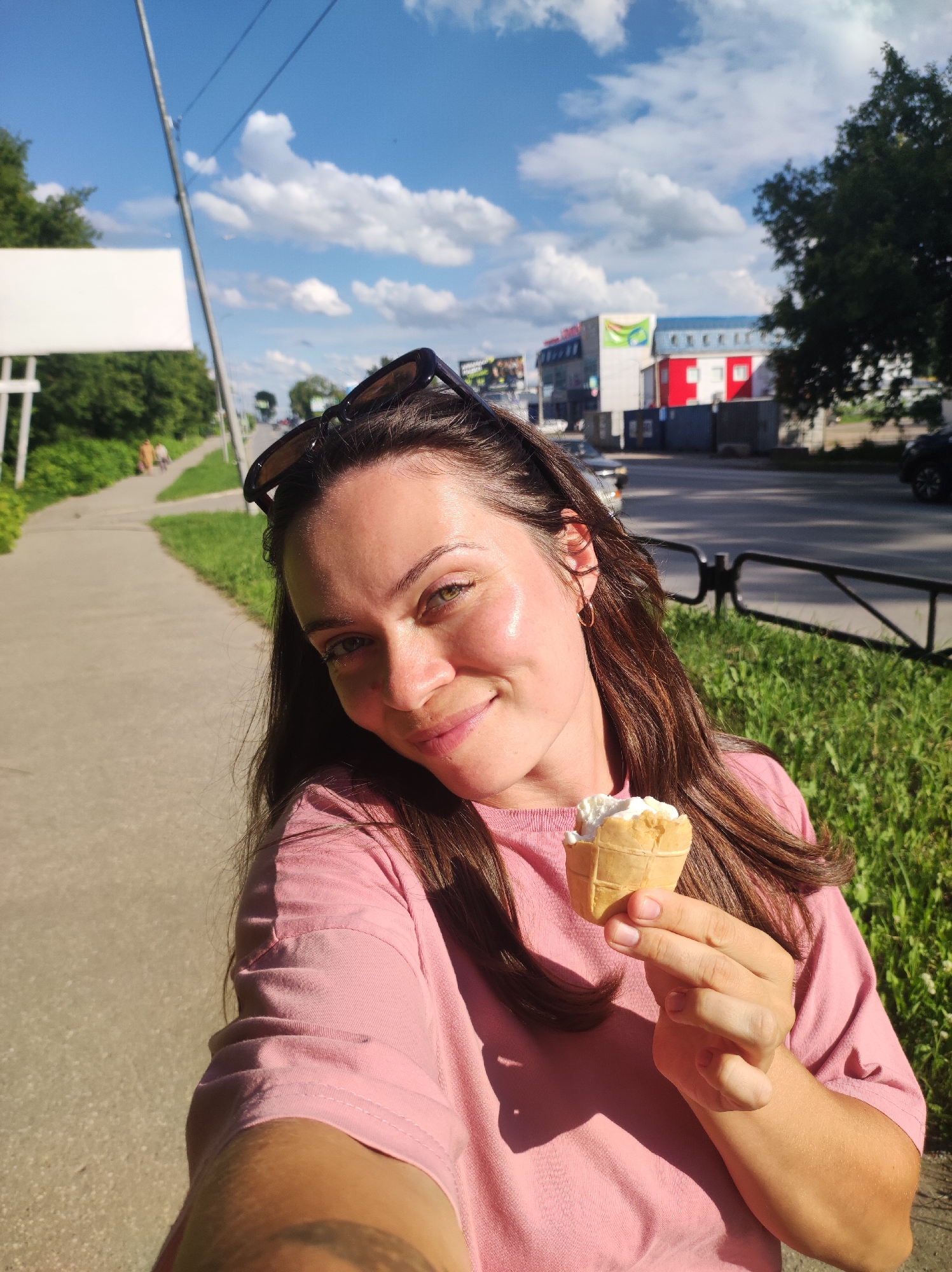 A woman is taking a selfie outdoors on a sunny day, holding an ice cream cone in her right hand. She is wearing a pink t-shirt and sunglasses are perched on her head. The background shows a sidewalk, a grassy area, trees, a road with cars, and buildings. The scene is likely located in the city of Chaikovsky, Russia, given the context. The woman appears to be smiling.