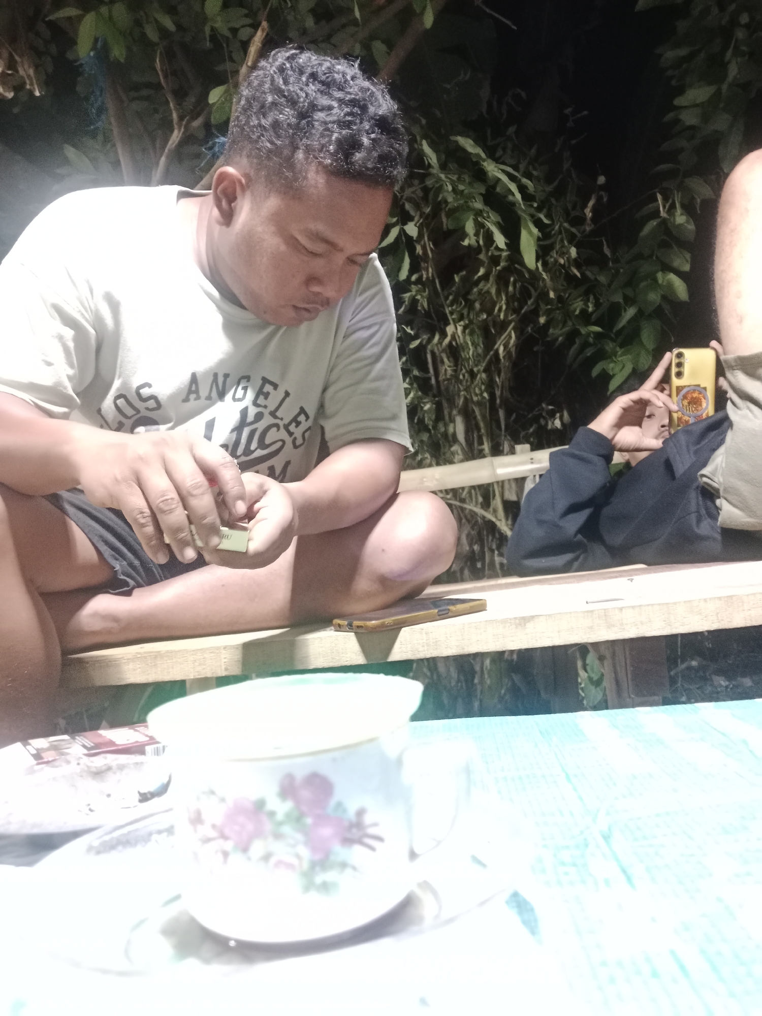 The image depicts a scene in Glagah Wangi, Indonesia, focusing on a man sitting outdoors, possibly at night given the lighting. The man is wearing a light-colored t-shirt with "LOS ANGELES" and "TIC" visible on it, and dark shorts. He is seated on a wooden bench and is holding a small, light-colored box, possibly cigarettes, in his hands. His gaze is directed downwards at the box, and his fingers are manipulating its contents. In the foreground, a white mug with a floral pattern is visible on a saucer, and next to it is a red and white pack of cigarettes. To the right of the man, another person is partially visible, holding a yellow smartphone, seemingly taking a selfie or recording. The background consists of dark foliage and trees, suggesting an outdoor, natural environment. The overall impression is a casual, relaxed gathering.