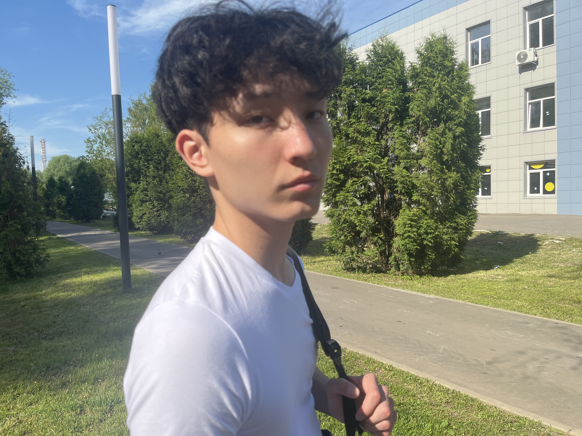 The location is the city of Unknown, Unknown.
A young man stands outdoors, angled slightly to his left, looking directly at the camera. He has dark, wavy hair and wears a white V-neck t-shirt. A black strap, likely from a bag, passes over his left shoulder and is held by his right hand. The lighting indicates bright sunlight, casting shadows on the right side of his face and parts of the ground.
He is positioned on the edge of a light grey asphalt path that features several indistinct white chalk markings. Green grass verges line both sides of the path. To the man's left, beyond the path, are dense green shrubs and trees, followed by two tall, slender street light poles, one with a white top and black base, and another further in the distance. In the far background, against a blue sky with scattered white clouds, faint industrial structures are visible.
To the man's right and extending into the background is a multi-story building with a facade composed of light grey tiles and blue accents along the top edge and right side. It has multiple rectangular windows with white frames. An exterior air conditioning unit is mounted on the facade below a window. The lowest visible windows on the right side feature distinctive yellow circular and white irregular shapes affixed to the glass. Tall, dense green coniferous trees stand between the path and the building on the right side of the frame.