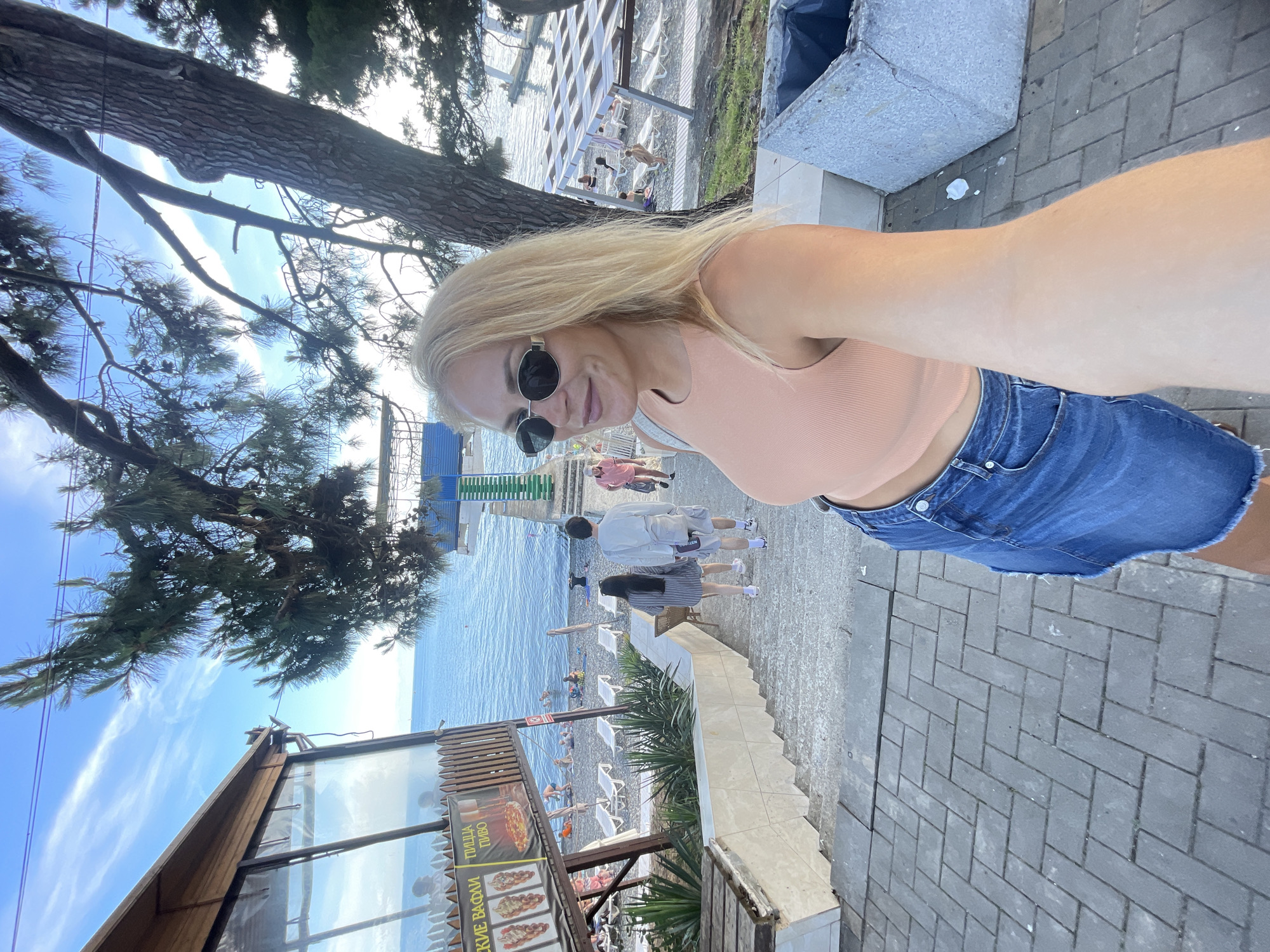 In Sochi, Russia, a blonde woman in her 30s, wearing an orange crop top and a denim skirt, takes a selfie on a paved pathway with a stone planter to her right. She is smiling, wearing sunglasses, and has her left arm extended. Behind her, a set of stone stairs descends towards a pebble beach. A couple, a man and woman, are walking down the stairs away from the camera. The beach is dotted with sun loungers, some occupied by beachgoers. A pier extends into the calm, blue sea, with a building housing a cafe visible on the left side of the frame, displaying signage for waffles and pizza. Lush green trees with visible bark frame the scene, with branches extending over the beach and sea, suggesting a seaside park or resort area. The sky is a clear blue with wispy white clouds.