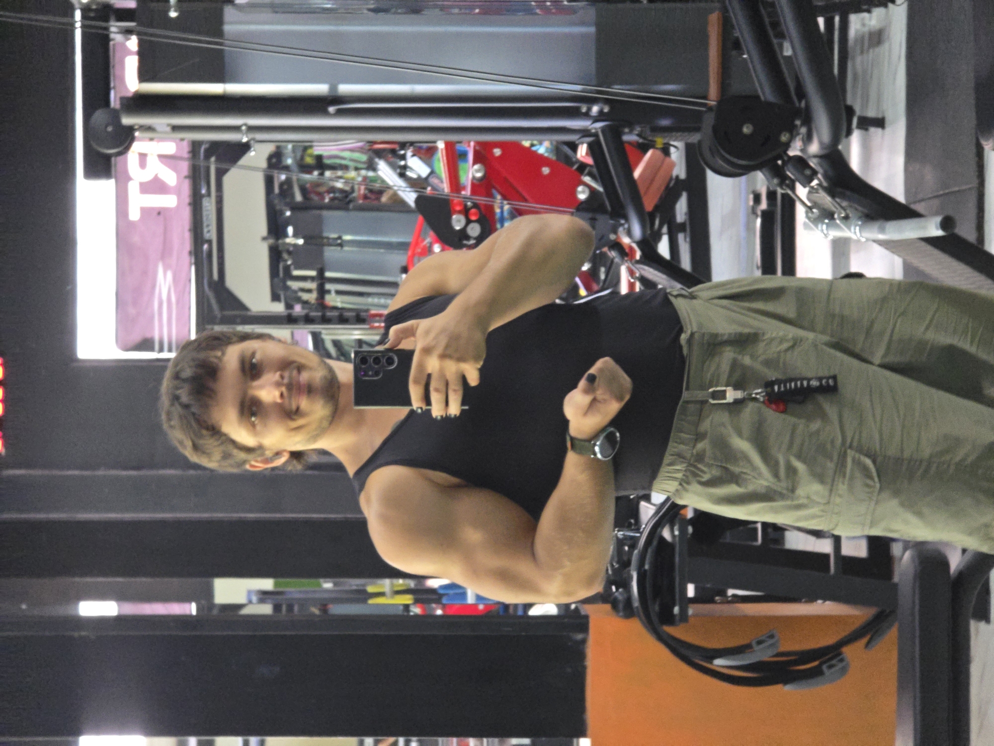 A man is taking a selfie in what appears to be a gym. The man has short brown hair and a beard. He is wearing a black tank top and khaki pants. He is holding a black smartphone in his left hand and flexing his right bicep. He is also wearing a watch on his right wrist. In the background, there are various pieces of gym equipment, including a cable machine and weight racks. A red sign with white text is visible in the upper left corner. The photo might have been taken in North Kuta, Indonesia.