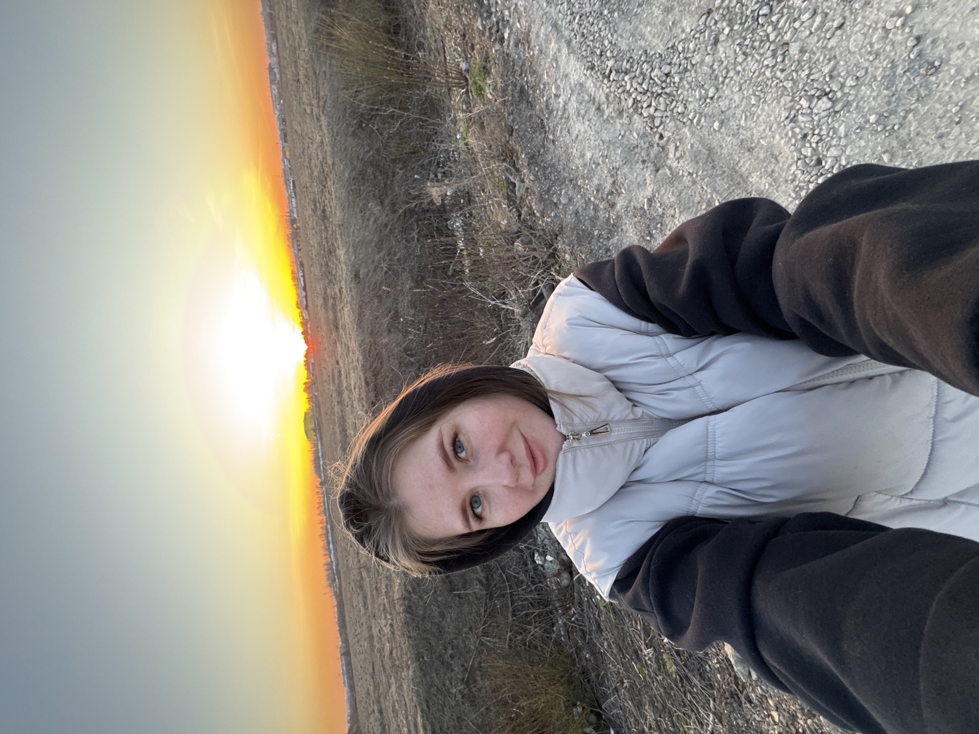 The image shows a young woman taking a selfie outdoors at what appears to be sunset. She is wearing a white puffer vest over a black hooded sweatshirt. The background features a field or open area with dry grass and a gravel or dirt road. The sun is setting on the horizon, casting a warm, golden light across the sky. The city of Горный is not visually discernible in the image.