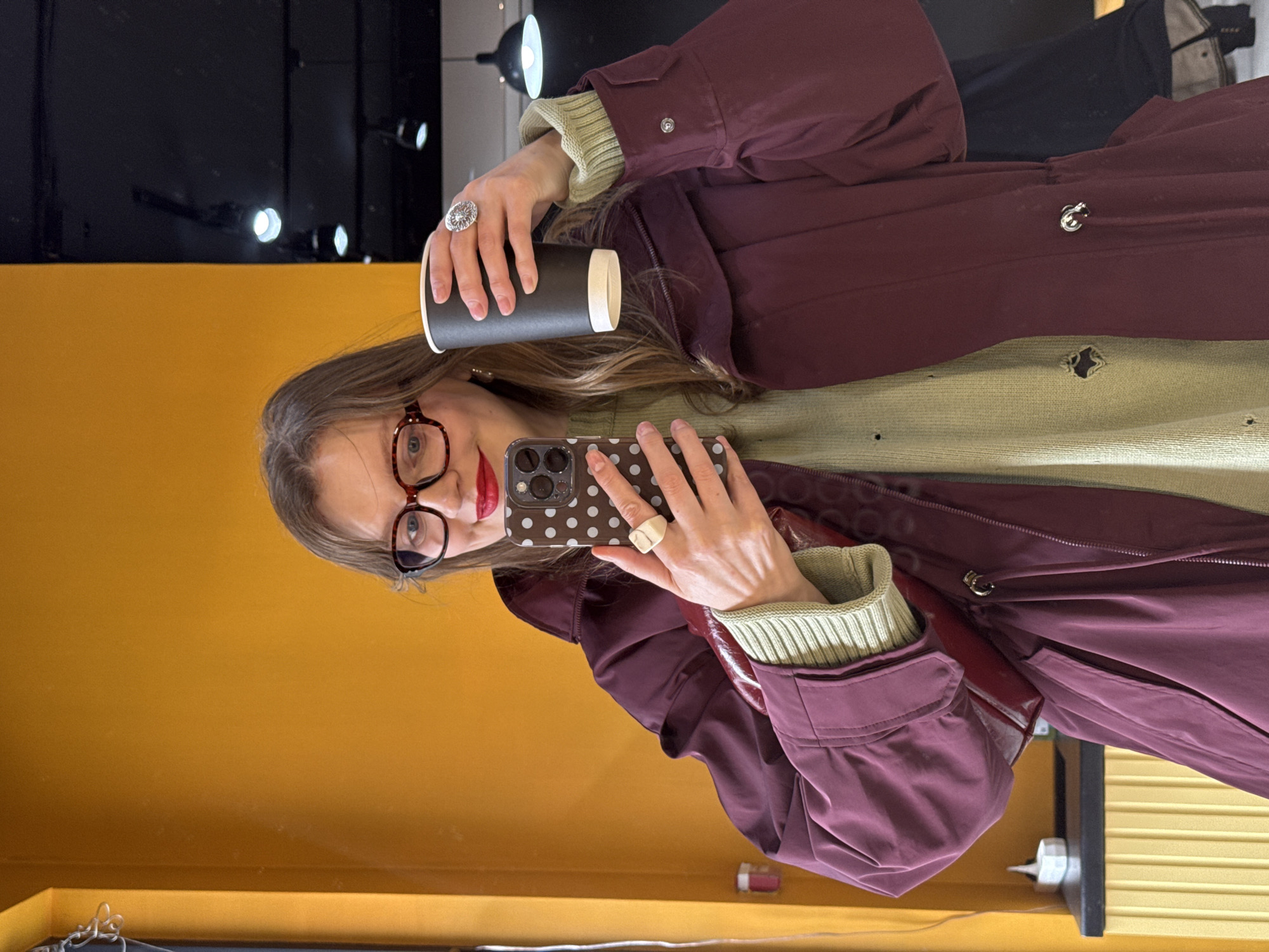 A woman with red lipstick and tortoiseshell glasses is reflected in a mirror, holding a black coffee cup in her left hand and a smartphone with a polka-dot case in her right hand. She wears a light green sweater and a burgundy jacket. The reflection shows a mustard yellow wall below the mirror, and above, black fixtures with spotlights, possibly in Izhevsk, Russia.