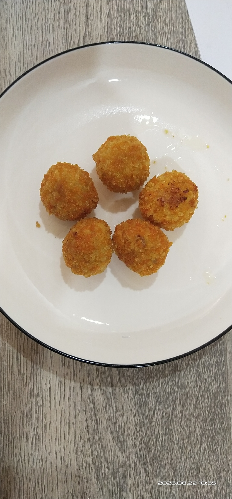 Five golden-brown, breaded croquettes are arranged in a circular pattern on a white plate with a black rim, which is placed on a wooden surface. The plate is slightly tilted in the frame, with the wooden surface extending to the bottom and left of the image. There are a few small specks of what appears to be sauce or oil on the plate. The lighting is overhead, casting subtle shadows. The scene is indoors and shows a close-up view of the food. The image is dated "2026.08.22 10:55" in the bottom right corner. This image does not contain information about a specific location in Matabu, Indonesia.