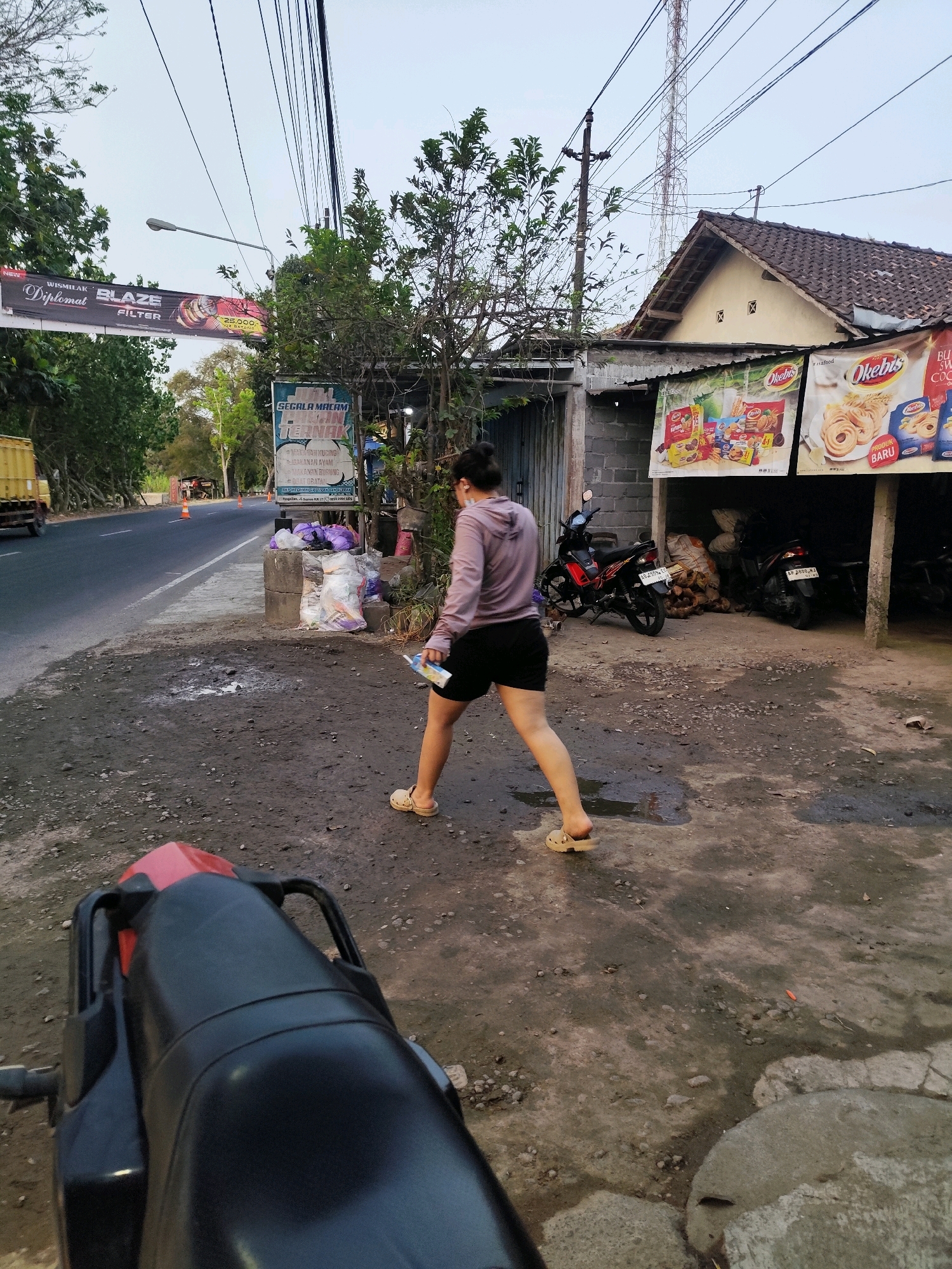 A young woman, wearing a light purple hooded sweatshirt and black shorts, walks away from the camera on a dusty, unpaved road. She is wearing light-colored clogs and holding a small white object, possibly a packet or a box, in her right hand. The scene appears to be on the side of a rural road in Bambanglipuro, Indonesia, indicated by signage in Indonesian.
To the left of the woman, a paved road stretches into the distance, with trees lining its edges and a yellow truck visible in the background. A banner advertising "Wismilak Diplomat Blaze Filter" is suspended over the road. To the right of the woman, a small building with a tiled roof is visible. Advertising posters for "Okebis" food products are attached to the front of the building, which seems to be a small shop or warung. Two motorcycles are parked near the building, one black and red, the other black. Bags of what appear to be coconuts are stacked in the open doorway. The ground is a mixture of dirt, gravel, and some puddles, suggesting recent rain. Utility poles and power lines are prominent in the sky. The overall atmosphere is that of a typical roadside scene in a semi-urban or rural Indonesian setting.