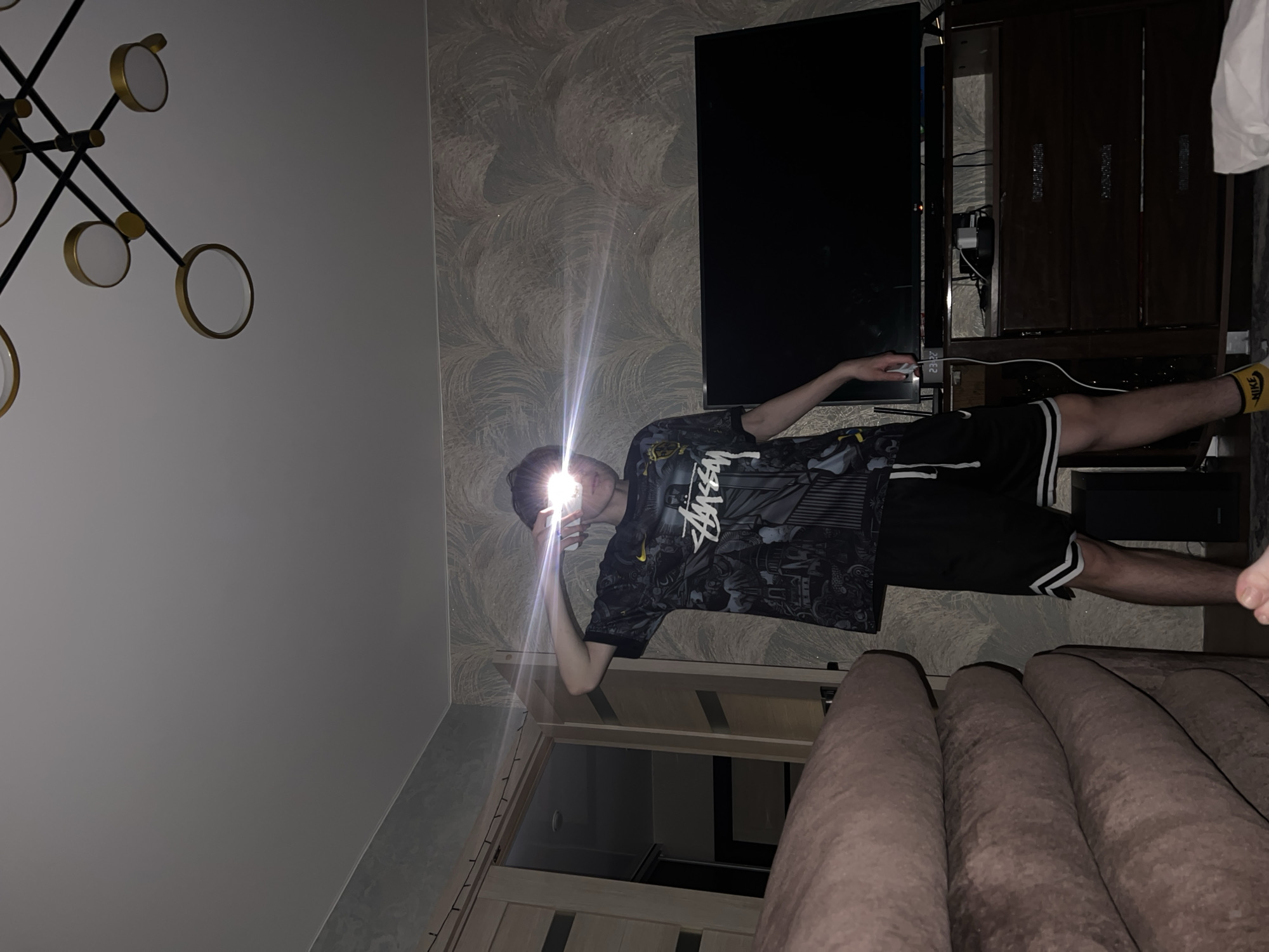 A person stands in a room, holding a phone with its flashlight on, obscuring their face with a bright light. They wear a patterned black t-shirt and black shorts with white stripes. Behind them is a patterned wallpaper, a large television, and a dark-colored cabinet. The room also features a modern chandelier with circular lights and a brown couch. The city of Noviy, Russia cannot be determined from the image.