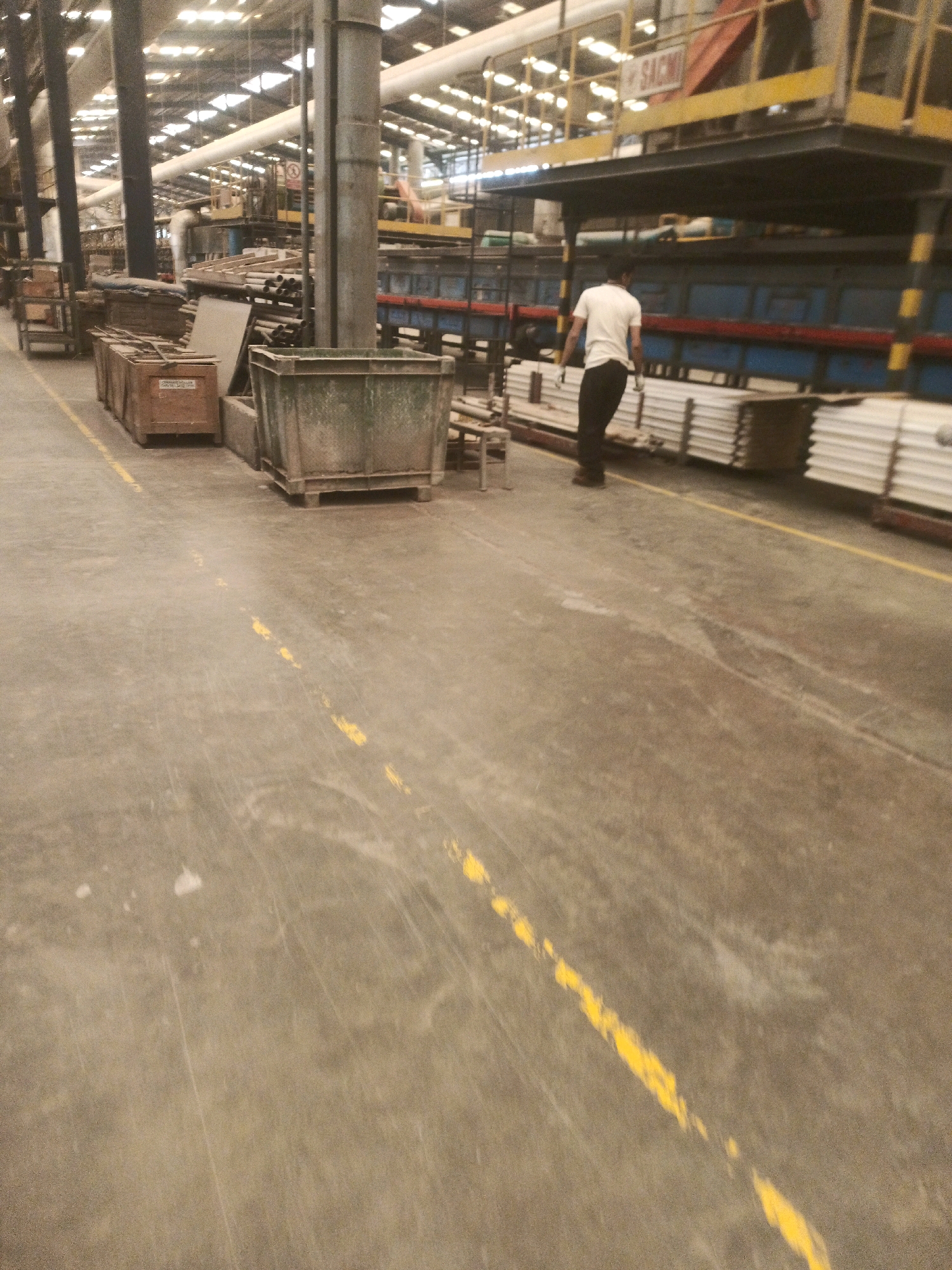 A man in a white t-shirt and black pants walks down a factory aisle in Lebaniwaras, Indonesia. The aisle is marked with yellow lines. Pallets of white materials are stacked to the right of the man. To the left, there are several wooden crates and a large, green, metal bin. The factory appears to be a production facility with industrial machinery and overhead pipes visible in the background. The lighting is bright, suggesting artificial lighting common in industrial settings.
