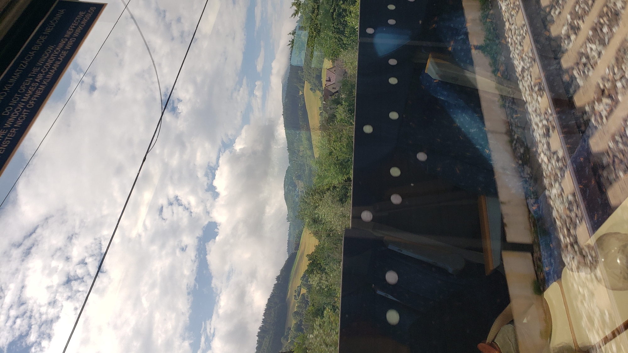 The image shows a view from a moving train, likely in a rural area. In the foreground, there are train tracks and a dark barrier with circular reflections. Beyond the barrier, a lush green landscape with rolling hills and scattered trees is visible. Some houses are nestled among the trees on a hillside. The sky is partly cloudy with bright sunlight. A sign above the window reads, "DO NOT OPEN THE WINDOW, THE WINDOW MAKES AIR-CONDITIONING INEFFECTIVE. ENSTER NICHT OFFNEM, KLIMAANLAGE WIRD WIRKUNGSLOS." The text suggests the train is equipped with air conditioning and opening the window would disrupt its effectiveness. Based on the language on the sign and the general scenery, the location is likely in a Central European country. Although specific details to pinpoint Maršová-Rašov, Slovakia are not definitively present, the landscape is consistent with rural areas in Slovakia.