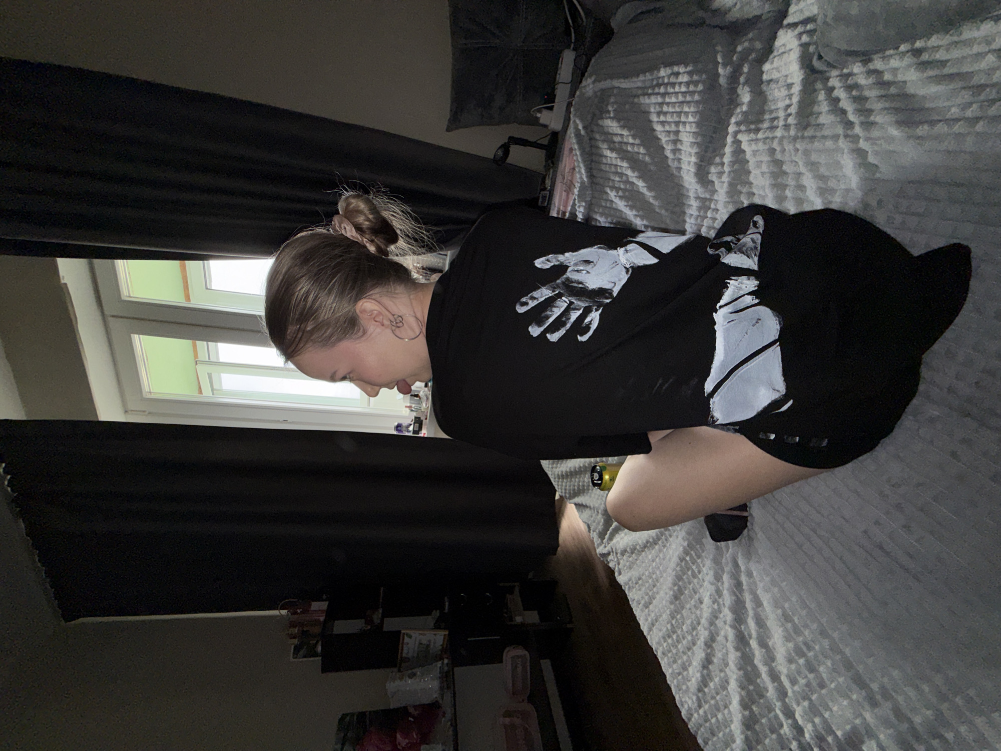 The image depicts a person kneeling or crouching on a bed in an indoor setting, likely a bedroom in Chaikovsky, Russia. The subject, a young individual with light brown hair tied in a bun, is viewed from a side profile, bending forward and looking downwards. They are wearing a dark, long-sleeved shirt featuring a large white graphic of an open hand on the back. Dark shorts and socks are also visible, along with a silver hoop earring in the ear.
The bed is covered with a light-colored, textured blanket. Heavy, dark curtains are drawn across a window, partially revealing a bright window frame and a glimpse of green outside, suggesting daylight, although the room itself appears dimly lit. In the background, a wooden floor is visible, along with shelves or furniture holding various indistinct objects. A pillow and an extension cord are also discernible near the headboard. The person's posture suggests a moment of focus or contemplation. No explicit text is visible beyond the hand graphic on the shirt.