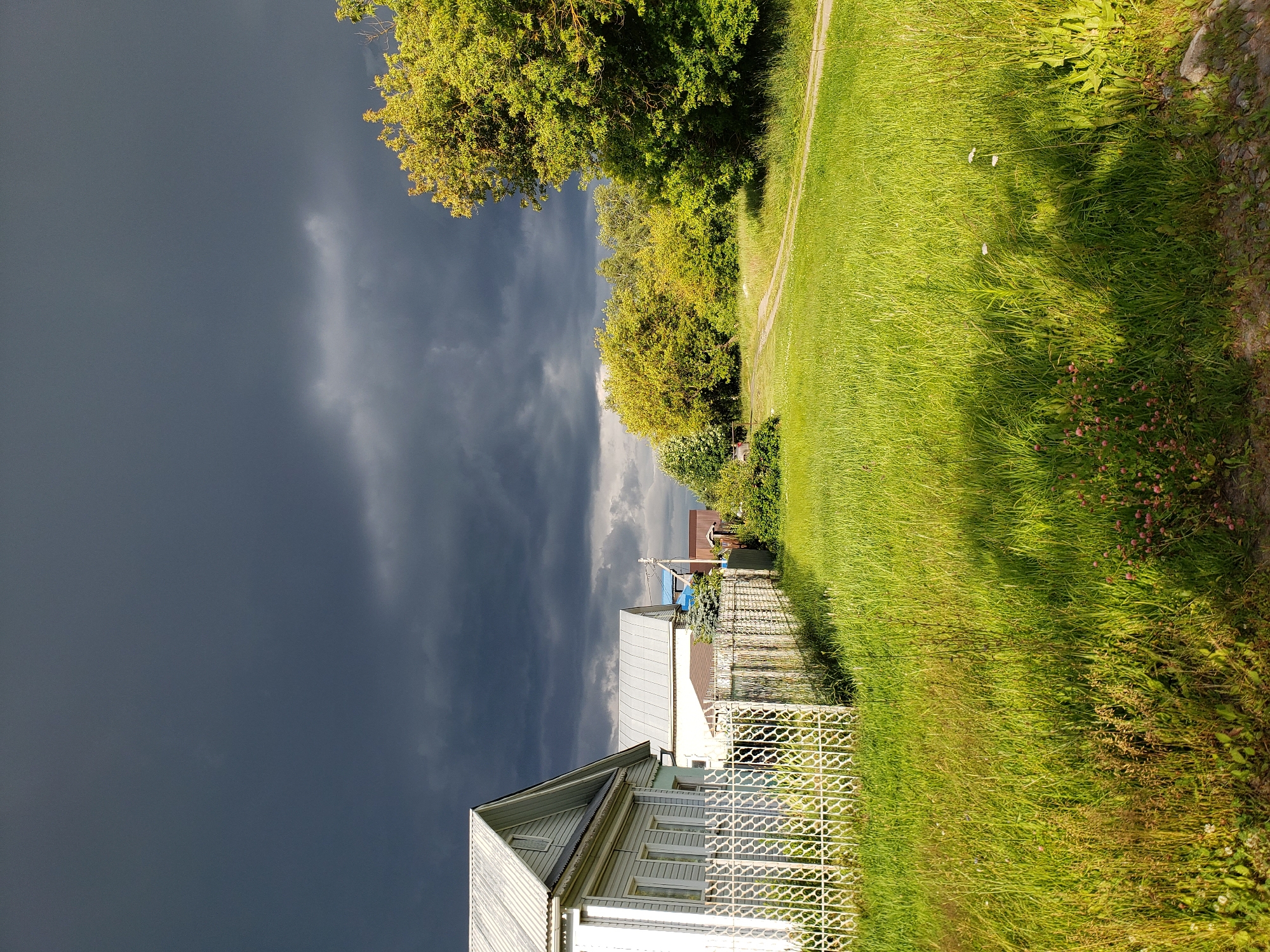 This media file captures a rural scene under a dramatic, overcast sky, suggesting an approaching storm. The sky is a deep, ominous grey, a stark contrast to the vibrant green of the well-lit grass and trees. In the foreground, a light-colored wooden house with a corrugated metal roof is partially visible, extending towards the right edge of the frame. A decorative white metal fence runs along the front of the property.
Behind the house, several other structures are visible, including another house with a lighter roof and a glimpse of red roofing material further back. The landscape is dominated by lush green grass and dense foliage from trees. A dirt path, or a narrow road, can be seen winding through the grass, leading away from the houses and towards the trees. The setting appears to be a village or a suburban area with houses and yards. The lighting suggests it is daytime, though the dark clouds create a moody atmosphere. The image evokes a sense of calm before a storm, with the natural elements of the weather playing a significant role in the scene's mood. There are no visible people in the frame. No text is visible. The location is identified as Nizhny Novgorod, Russia.