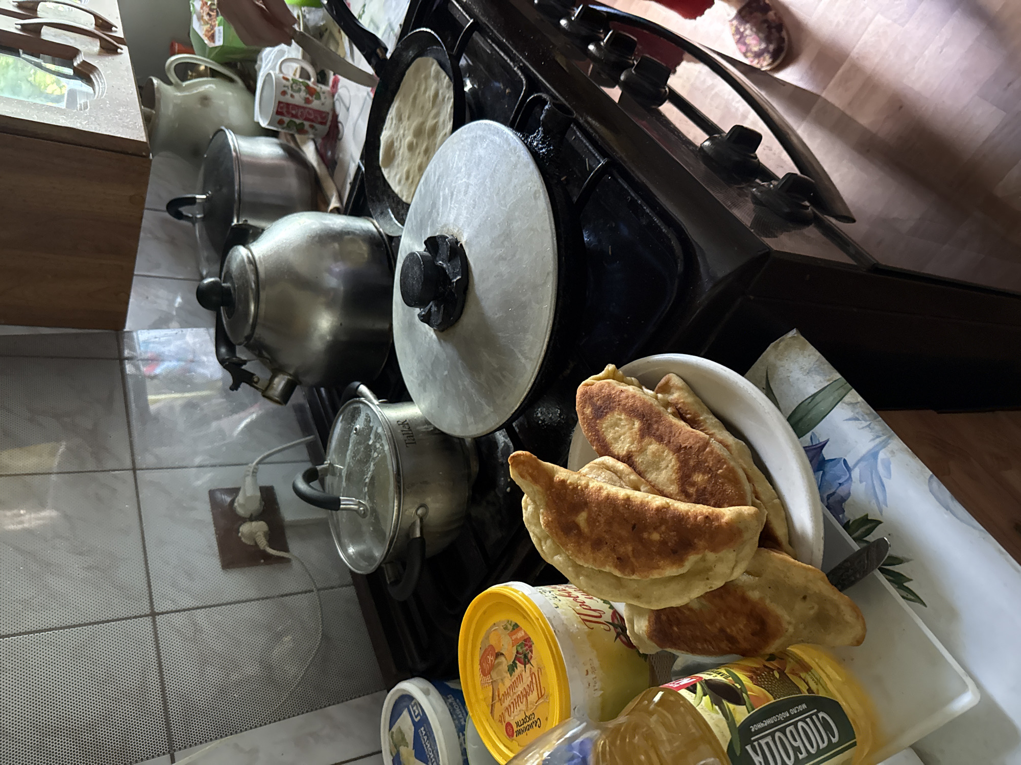 This image depicts a close-up view of a gas stove in a kitchen, likely in Valday, Russia. Several cooking items are present. On the stovetop, a metal teapot, a covered pot, and two pans are visible. One pan contains a flatbread, and another is covered with a large metal lid. A plate of fried pastries, possibly chebureki or pirozhki, is situated on the left side of the stove. Next to the plate, a bottle of sunflower oil with a label in Russian is placed on a white tray. Two tubs of dairy products are also on the counter to the left. The kitchen backsplash is tiled with a light-colored pattern. In the background, a wooden cabinet is visible. A person's hand is seen in the upper right corner, holding a knife, suggesting activity in the kitchen. The floor appears to be light-colored wood.