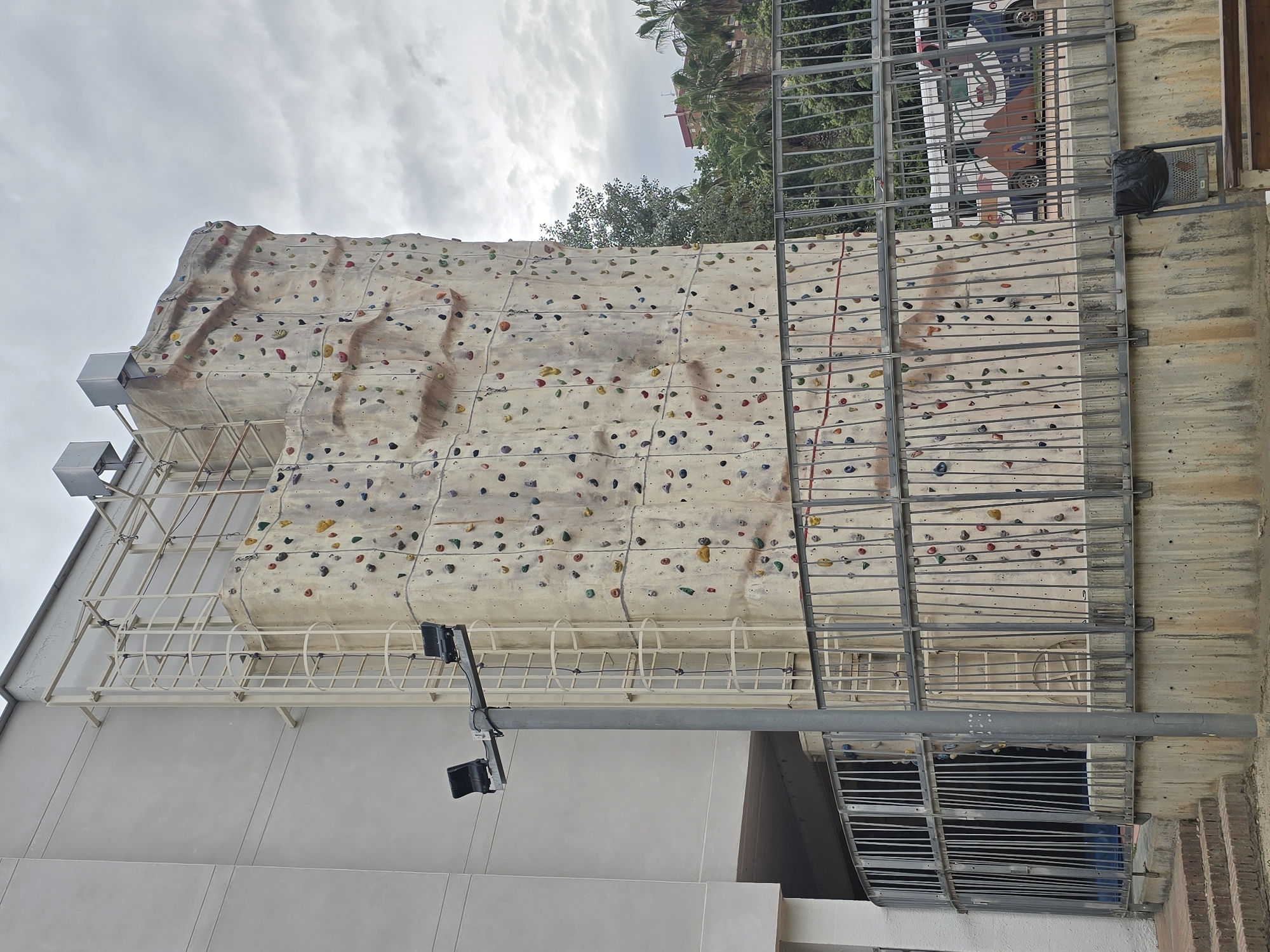 The image depicts an outdoor climbing wall situated in Cornellà de Llobregat, Spain. The wall is multi-paneled and has various colorful climbing holds attached. The wall's facade is painted in light shades, mimicking a natural rock surface. A metal scaffolding framework surrounds the climbing wall, providing structural support and safety measures. The climbing wall is attached to the side of a multi-story building with a pale-colored facade. The surroundings show trees and other infrastructure suggesting an urban environment. The sky is overcast, providing soft light on the objects in the scene. A streetlight with dual lamps is mounted near the bottom part of the image.