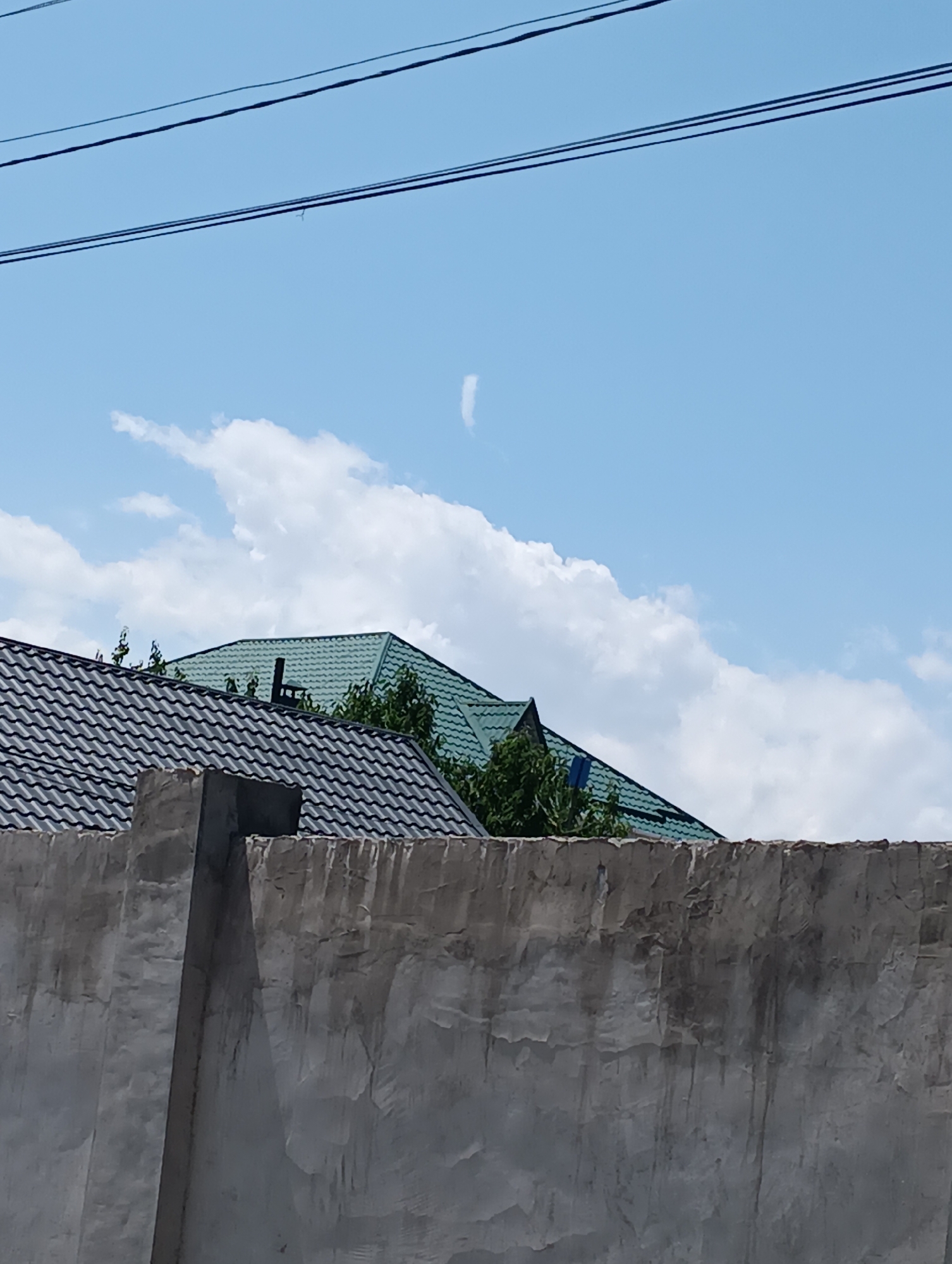 The image shows a close-up view of a textured concrete wall in the foreground. Behind the wall, two rooftops are visible, one with dark grey shingles and another with green metal roofing. Trees partially obscure the green roof. The sky above is a clear blue with scattered white clouds. Three dark electrical wires are strung across the top of the frame. No people are visible in the image. The scene appears to be an outdoor urban or suburban environment, possibly in Bishkek City, Kyrgyzstan, given the prompt.