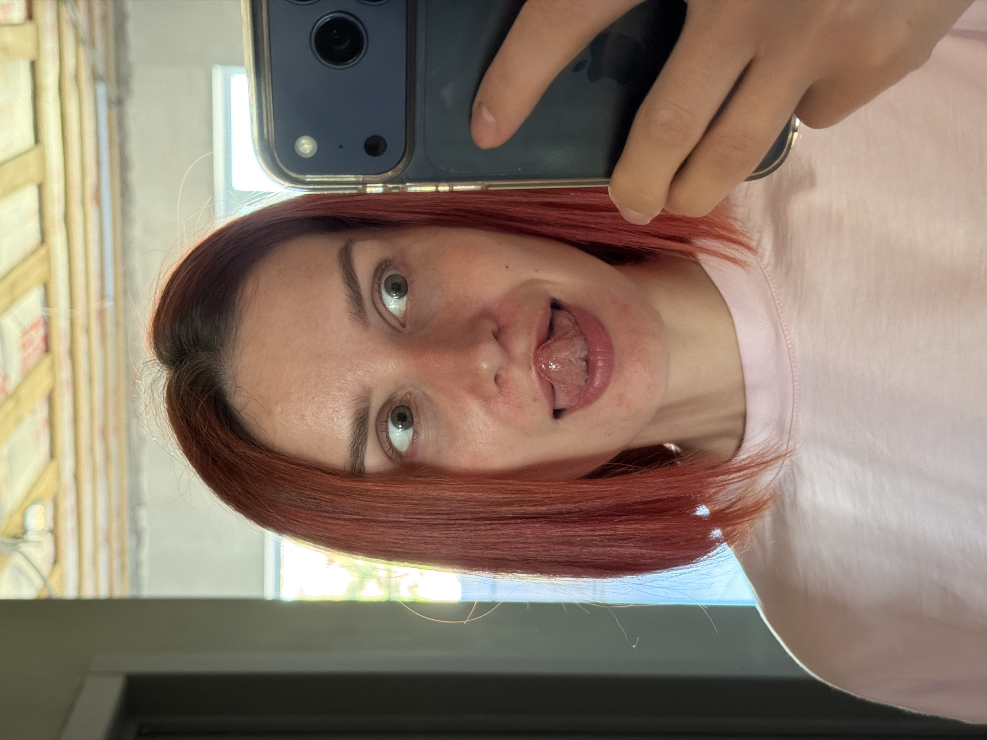 A light-skinned female with red hair takes a selfie with her phone. The woman is wearing a pink t-shirt. She is sticking her tongue out towards the camera. In the background, a window is visible, and a blurred object could be the building structure, potentially located in the city of Chaikovsky, Russia.