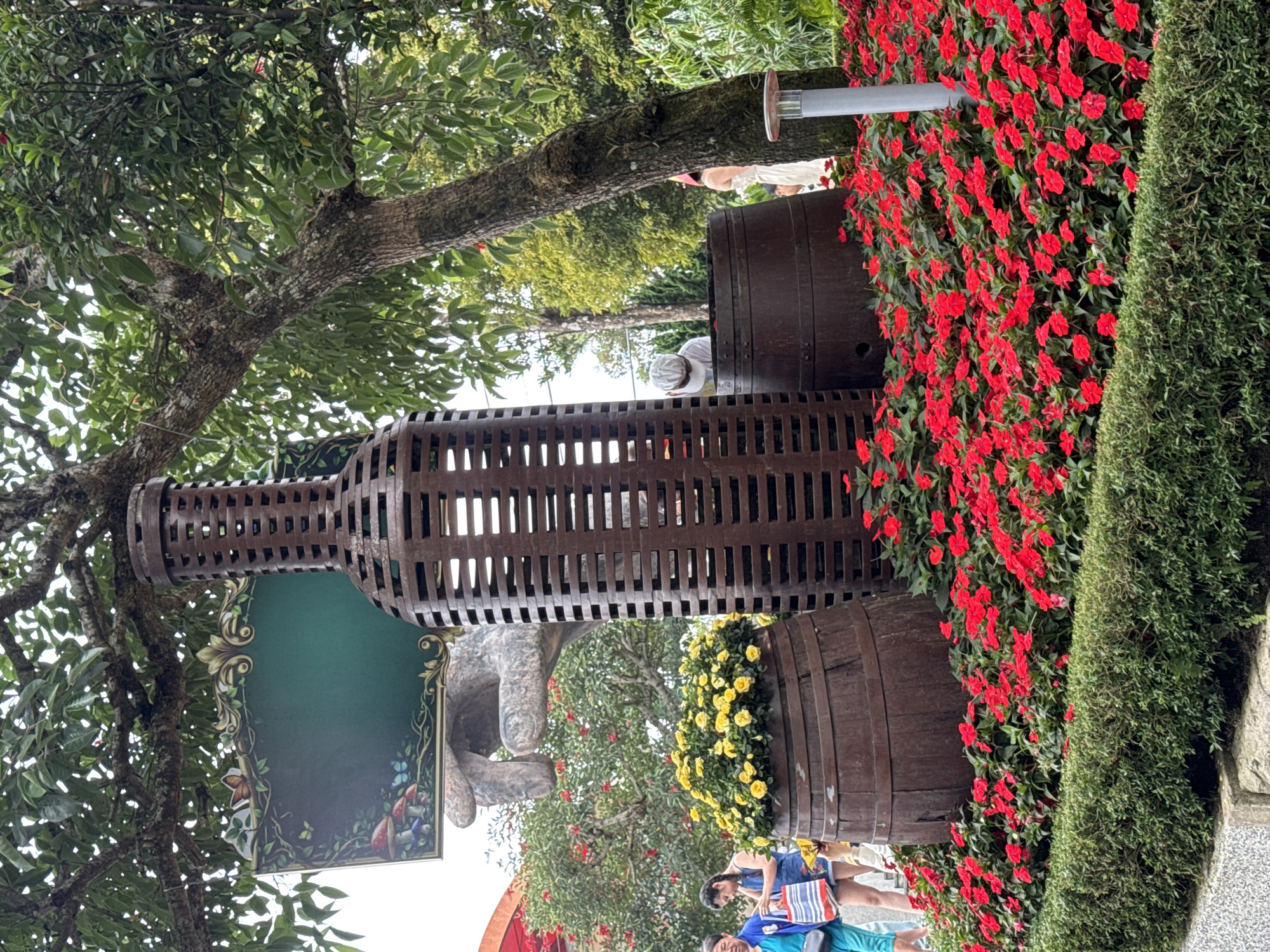 The image presents a scene at the Ba Na Hills resort, a tourist destination in Đà Nẵng, Vietnam. The focal point is a large, decorative sculpture resembling a wine bottle crafted from brown, latticed metal. The sculpture is positioned above two wooden barrels, each serving as a planter. The upper barrel is adorned with vibrant red flowers. The lower barrel is filled with bright yellow blooms.
Adjacent to the barrels, on the right side of the image, is a hedge of dense green foliage. The sculpture is set against a backdrop of trees with green leaves. To the left of the bottle sculpture is a large sign featuring painted decorations around a blank field.
In the lower portion of the image, there are two people, at least one woman, present in the scene, suggesting that the photograph was taken in a public area frequented by visitors.