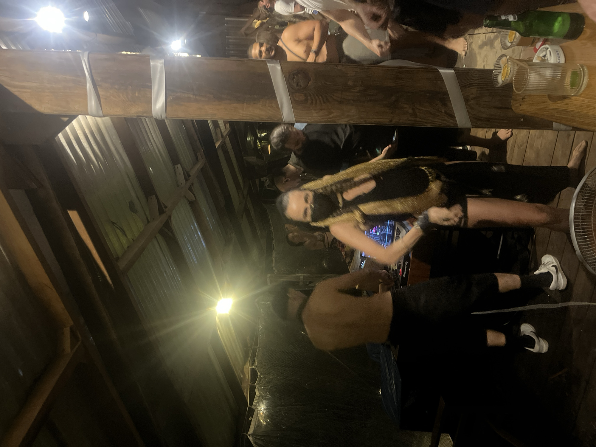 The image depicts an outdoor gathering at what appears to be a bar or club in Đà Nẵng, Vietnam. A wooden pillar, wrapped with two white bands, stands prominently in the foreground. Several individuals are visible. On the left, a shirtless person with a dark hat is near a DJ setup, with a woman in a black outfit and a gold sash standing in front of them, facing the camera. To the right of the pillar, a man with a beard, shirtless and wearing light shorts, is visible. Other people are seen in the background, some dancing or socializing. The setting has a rustic, open-air feel, with corrugated metal roofing and wooden structures. Several glasses and a green bottle are on a wooden table in the lower right of the frame. The lighting suggests it is evening or night.