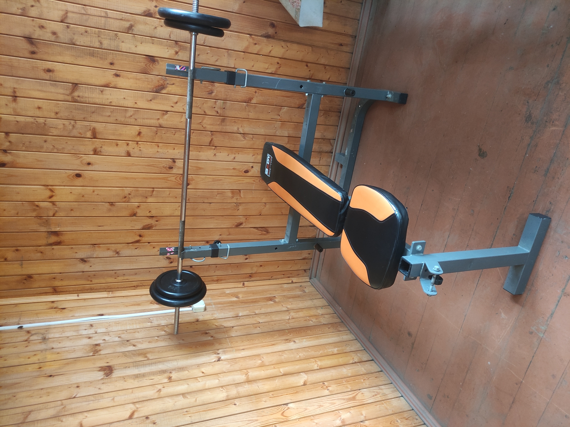 The image shows a weight bench and barbell stand with weights in a room with wooden paneling. The bench is gray with black and orange padding. The barbell is silver and has black weights on it. The room has wooden floors and walls. The location in Kratovo, Russia is not discernible from the image.