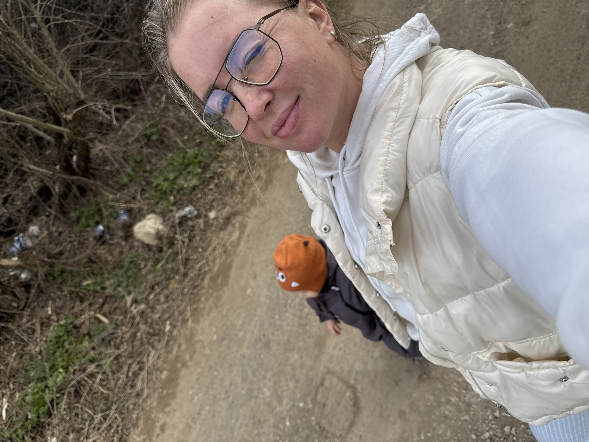 A woman wearing glasses and a white puffy jacket is taking a selfie. A child wearing an orange hat is visible below and to the left of the woman. They are on a dirt path with dry vegetation to the left, including trash. The location is likely Chaikovsky, Russia.