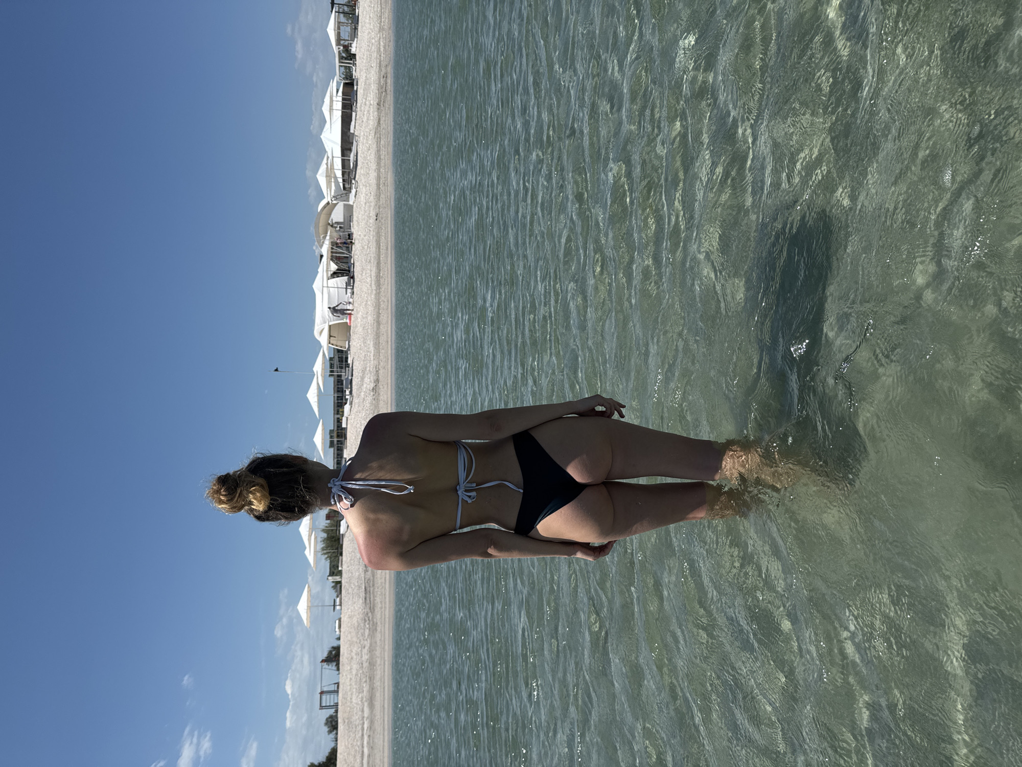 A medium, outdoor shot presents a woman wading in shallow, clear water at a beach in Мысовское сельское поселение, Ukraine. The woman, seen from behind, has a bun hairstyle and is wearing a two-piece swimsuit. The background features a sandy beach lined with white canopies, extending towards a clear blue sky. The water is slightly rippled, and her feet are partially submerged.