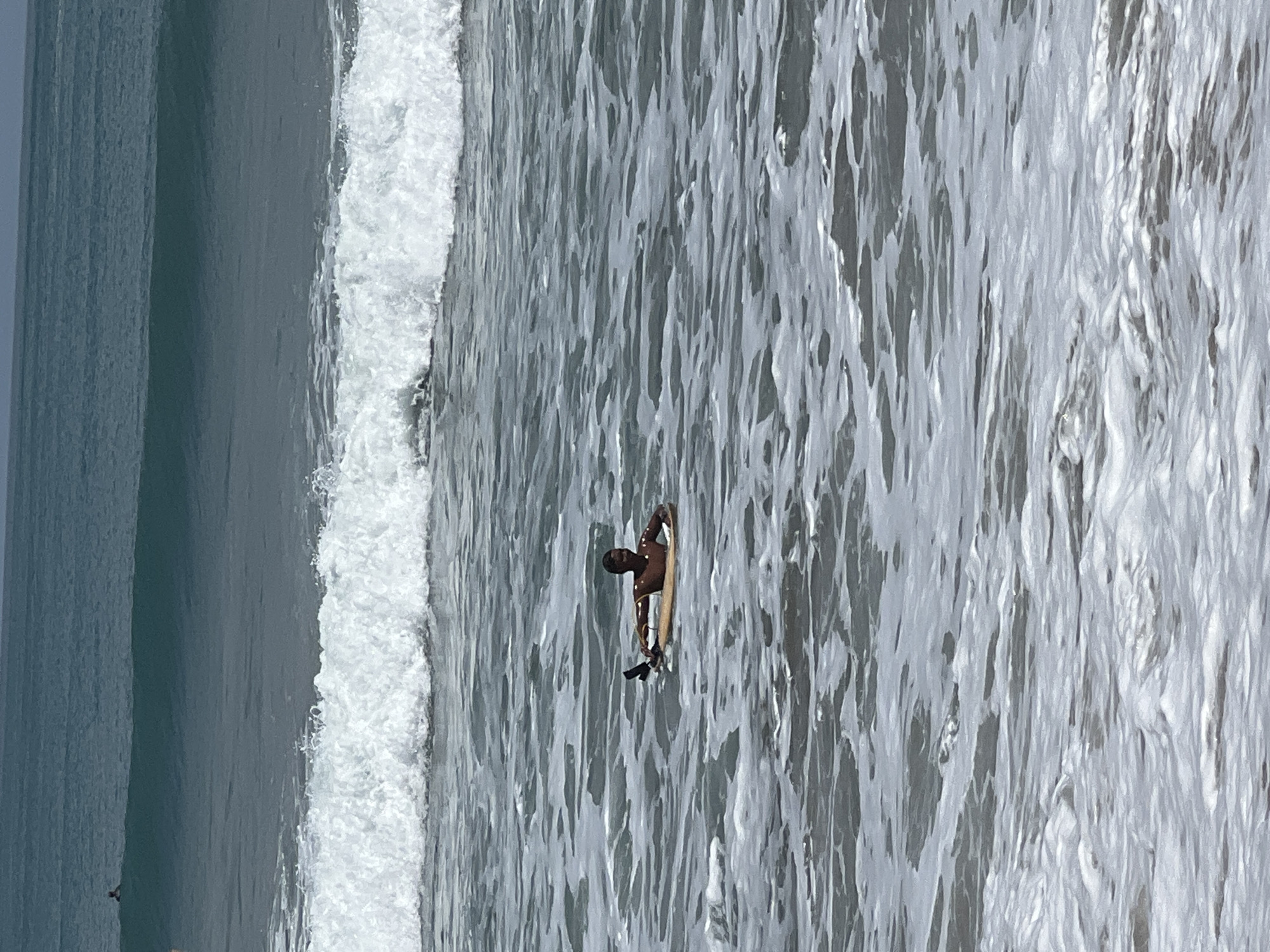 A person is floating on a boogie board in the ocean off Kuta, Indonesia. The person is positioned in the center of the frame, lying on their stomach on the board. The water is a muted blue-green, with visible ripples and some white foam. A wave is breaking in the background, forming a white crest. The horizon line is visible in the upper part of the image, separating the water from a pale sky. Another, smaller figure is visible further out in the ocean, appearing to be on a surfboard.