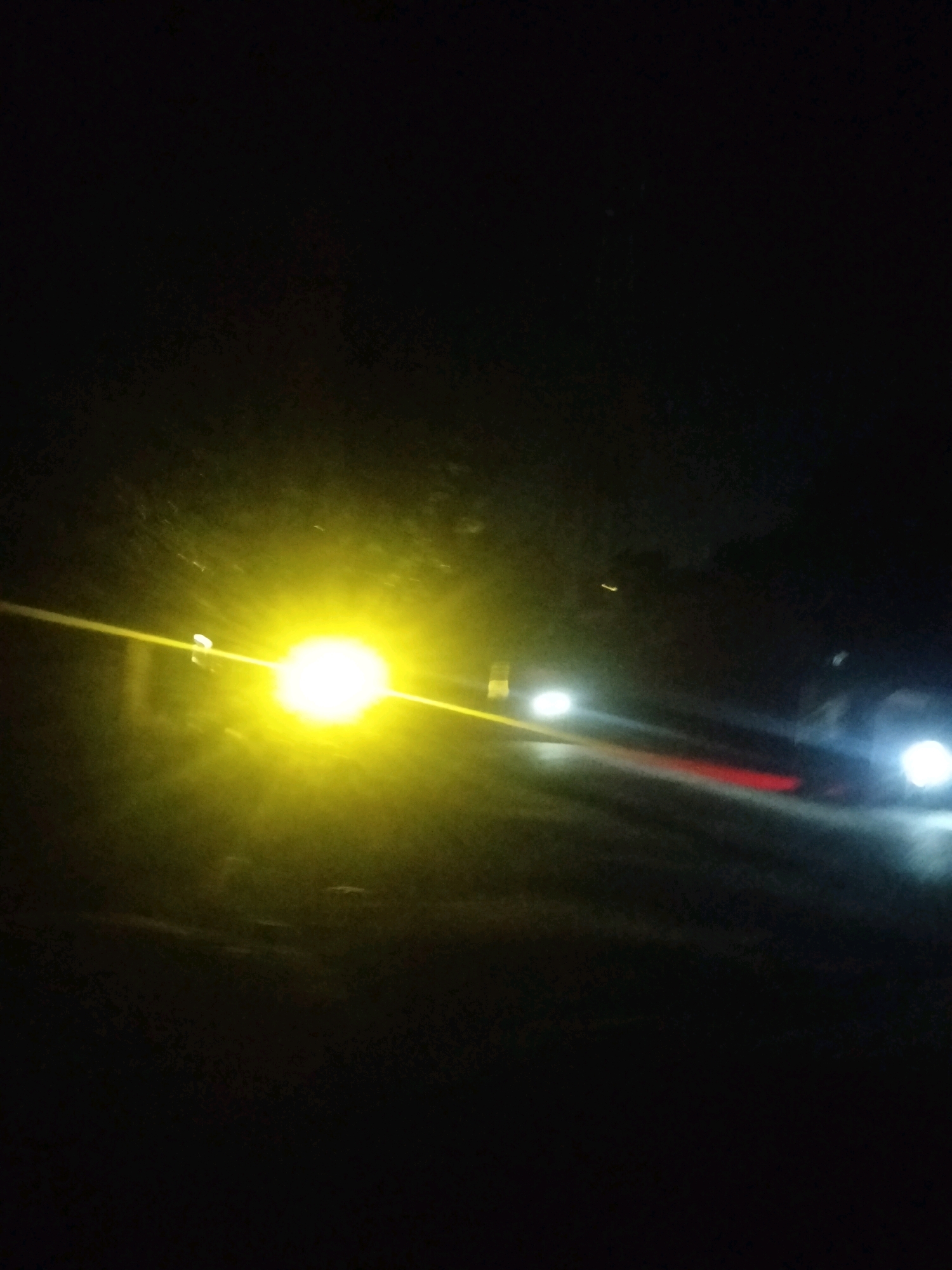 This image depicts a dark, nighttime scene in Bholagonj, Bangladesh, characterized by the presence of vehicle lights. A prominent, bright yellow light source dominates the left side of the frame, likely headlights or a powerful street lamp. To the right, other vehicle lights are visible, including white headlights and a distinct red taillight, suggesting vehicular movement on a road. The background is predominantly dark, with some faint shapes that could be trees or structures, hinting at the surrounding environment of Bholagonj. The overall impression is one of a dimly lit urban or semi-urban area at night, with traffic being the primary visual element.