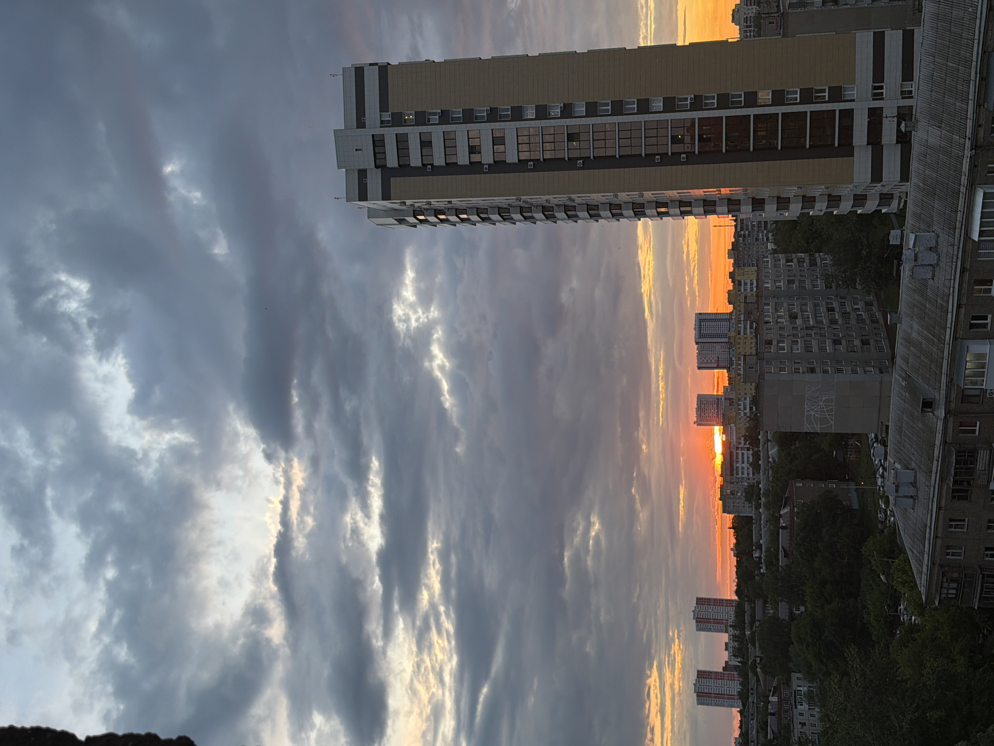 The image, correctly viewed horizontally, captures an urban skyline during a dramatic sunset. The sky is a prominent feature, dominated by dark, heavy grey clouds on the left and central portions, which transition into a brilliant expanse of orange and yellow light on the right. The sun, a vivid orange disc, is visible low on the horizon, partially obscured by distant buildings and clouds.
In the left foreground, a tall, modern high-rise building stands prominently. It features a light-colored facade with horizontal dark bands and numerous windows, some of which show reflections or internal lights. Several air conditioning units are mounted on its exterior.
The midground and background consist of a dense arrangement of residential buildings, including older, block-like structures with visible roofs and many windows. These buildings extend towards the horizon, interspersed with dark green trees. The scene conveys a sprawling city at the close of day, illuminated by the striking colors of the setting sun. No people or visible text are present.