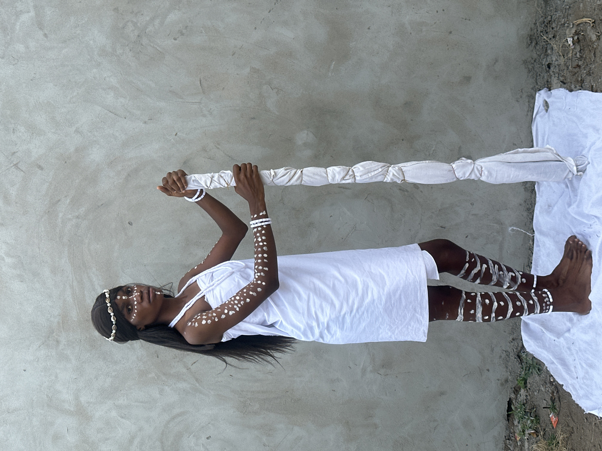 A young person stands in front of a textured concrete wall, dressed in a white garment and adorned with white body paint. They wear a headband made of shells and bracelets on both wrists. The body paint features abstract patterns on their face, arms, and legs. They are holding a long, white object wrapped in fabric, which appears to be a staff or pole. The ground beneath them is covered with a white cloth, and to the left, sparse vegetation is visible. The scene is located outdoors, likely in Badagry, Nigeria, given the cultural attire and context often associated with the region's traditional practices. The lighting suggests it is daytime.