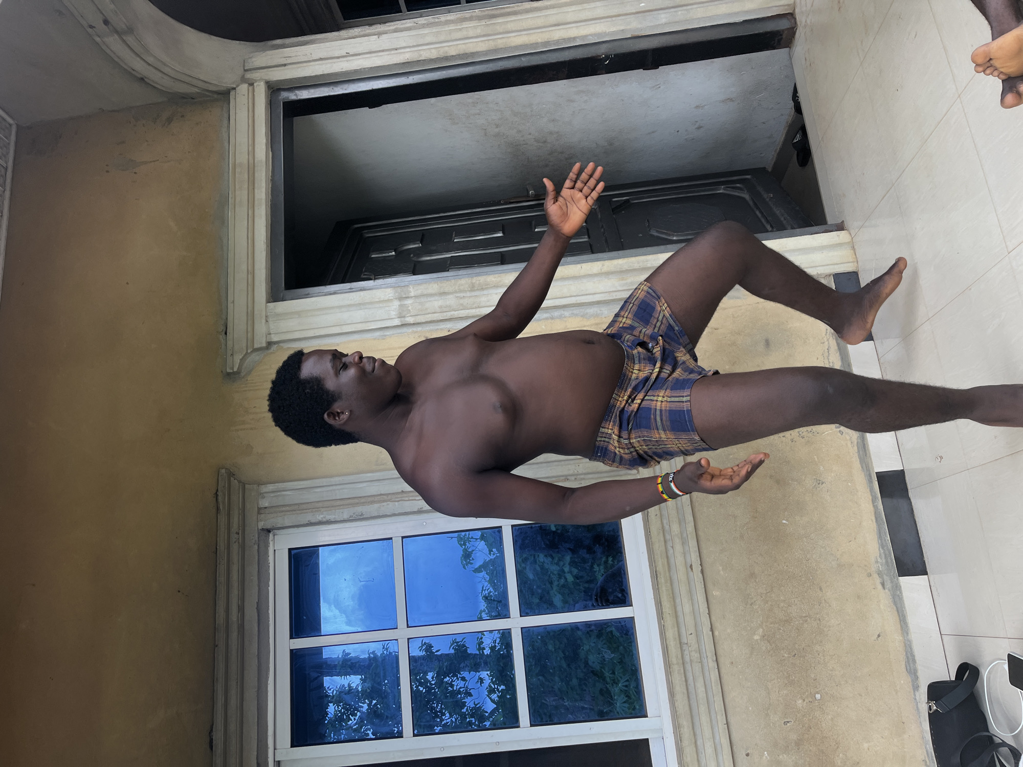 This vertically oriented image, if rotated counter-clockwise, depicts a shirtless dark-skinned man standing or leaning in what appears to be an outdoor or semi-outdoor setting, possibly a porch or veranda. He is wearing plaid shorts with blue, yellow, and brown squares. His head is tilted slightly back, and he looks upward with a neutral expression. Both his arms are raised, bent at the elbows, with open palms facing slightly upward, as if in a gesture of reception or offering. He has short, dark, curly hair, an earring in his left ear, and a multi-colored beaded bracelet on his right wrist.
The background features light brown walls with architectural moldings. To the man's left (our right in the rotated image), there is a white-framed window with multiple panes of blue-tinted glass, reflecting trees and possibly the sky. To his right (our left), a dark doorway or entrance with a dark door is visible. The floor beneath him is covered with light-colored tiles, possibly white, with some black squares visible in the distance. In the foreground on the tiled floor, a black bag and white cables are visible. A portion of another person's bare foot is visible at the very top right edge of the original image (top left after rotation), suggesting another individual is nearby.
The scene is brightly lit by natural light, indicating daytime. The overall ambiance is casual and relaxed, with the man seemingly posing for the photograph. There is no visible text in the image. The setting and subjects are consistent with the location context of Ikot Ekpene, Nigeria.
