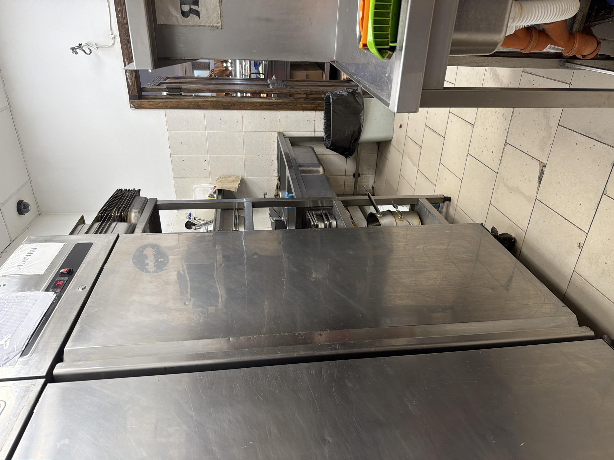 The media depicts a functional commercial kitchen or food preparation area in Plovdiv, Bulgaria. No individuals are present, and no active tasks are underway, suggesting a moment between operations or an overview of the workspace.
The scene is dominated by stainless steel equipment, including large refrigeration units or prep tables that show signs of regular use with scratches and water marks. One of these units features a digital display showing "0.6," likely indicating a temperature. On its surface, a paper label with partially visible Cyrillic text, possibly "ПЕЧЕНИ" (meaning "baked" or "roasted"), is under a plastic cover, suggesting menu or preparation information. A distinct "Batman" logo sticker is also affixed to the steel surface.
To the right, a stainless steel sink area is visible, with characteristic white corrugated drainage pipes and orange plumbing underneath. A black trash bag is placed in a white bin adjacent to the sink. The walls in this section are covered with light-colored tiles, and a wooden window frame reflects an internal space, possibly an adjacent room or service area.
The upper left portion of the image shows a white wall equipped with a small security camera and a simple light fixture. Below the main stainless steel units, additional shelving or equipment holds stacked kitchenware, possibly baking trays. The floor is tiled with light-colored, square tiles that exhibit some wear and smudges. The presence of Cyrillic text on the label reinforces the specified location context.