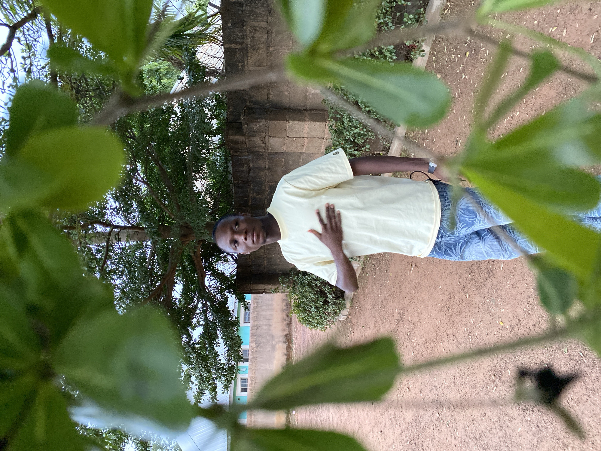 A young man stands in an outdoor area, likely a yard or garden, in what is presumed to be Bukuru, Nigeria. The man is light-skinned, wearing a light yellow shirt and blue patterned jeans. He has his right hand placed on his chest. The foreground is heavily obscured by green leaves, giving the impression that the picture was taken from behind or through foliage. Behind the man, there is a brick wall and green vegetation. A tree with thick branches is visible on the left side of the frame. In the background, there is a glimpse of a light blue and white building. The ground appears to be dirt.
