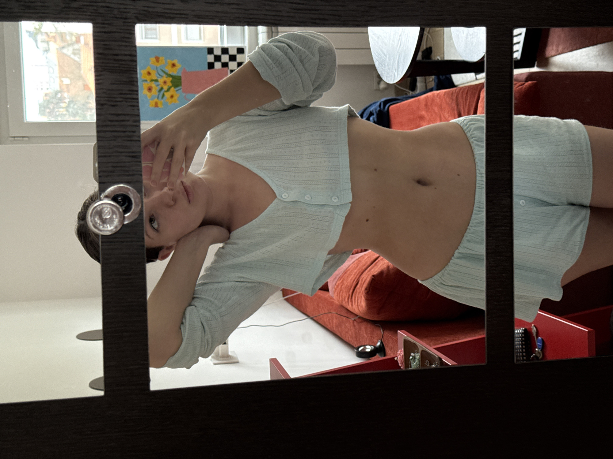 The image depicts a young adult female captured in a mirror selfie within an indoor room located in Vozdovac Urban Municipality, Serbia. She wears a light blue, textured two-piece loungewear set consisting of a cropped, button-front, V-neck top revealing her abdomen and matching shorts. Her right hand holds a light-colored smartphone, visible in the mirror, while her left arm is bent with the hand behind her head. The mirror features a dark frame and a circular door handle. Reflected in the background are white walls, a red upholstered sofa, and a multi-tiered red shelving unit displaying small decorative items and a set of keys with a blue fob. A round wooden table and a white radiator are also visible. On the wall, a framed artwork portrays yellow flowers in a pink pitcher on a black and white checkered surface. Through a window, residential buildings with red-tiled roofs and trees are visible, indicating an urban residential environment.