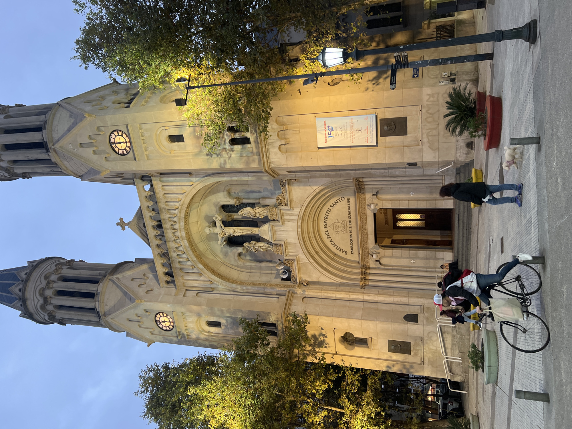 The image shows the Basilica del Espiritu Santo Parroquia N. S. de Guadalupe, a Catholic church in the city of Buenos Aires, Argentina. The facade of the church is visible, with a main entrance featuring statues and an inscription stating "BASILICA DEL ESPIRITU SANTO PARROQUIA N. S. DE GUADALUPE". The church has two towers with clocks. A streetlight illuminates the facade. On the right side, a woman walks a dog. A bicyclist is visible to the left of the door. A sign on the church says "AÑO JUBILAR VERBITA 150 TESTIGOS LUZ".