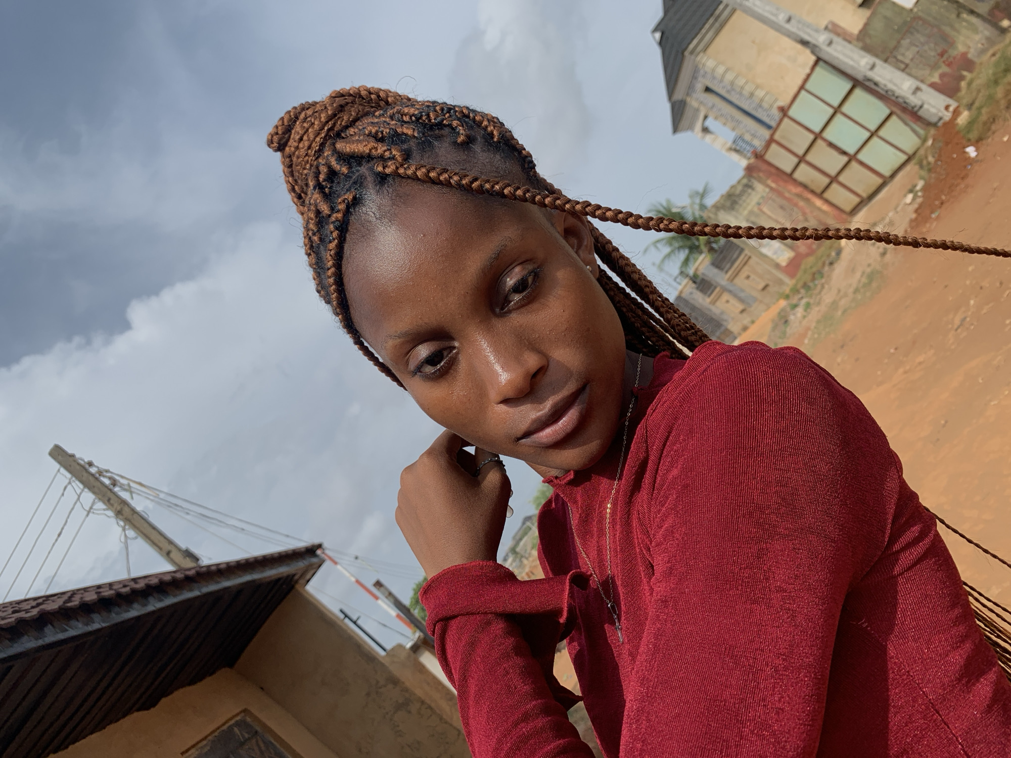 A medium-shot photograph shows a young, brown-skinned woman wearing a red, long-sleeved shirt, with her head slightly tilted down and to the left. Her hair is styled in neatly braided extensions, pulled up into a bun on top of her head, with several loose braids framing her face. She rests her chin on her left hand, and wears a necklace with a pendant.
In the background, the sky is overcast with clouds. Behind her and to the left, a dilapidated building can be seen, and a palm tree is visible behind it. The ground appears to be a dirt surface. In the lower left, there is another building with a corrugated roof and overhead power lines. This scene depicts a location in Ikorodu, Nigeria.
